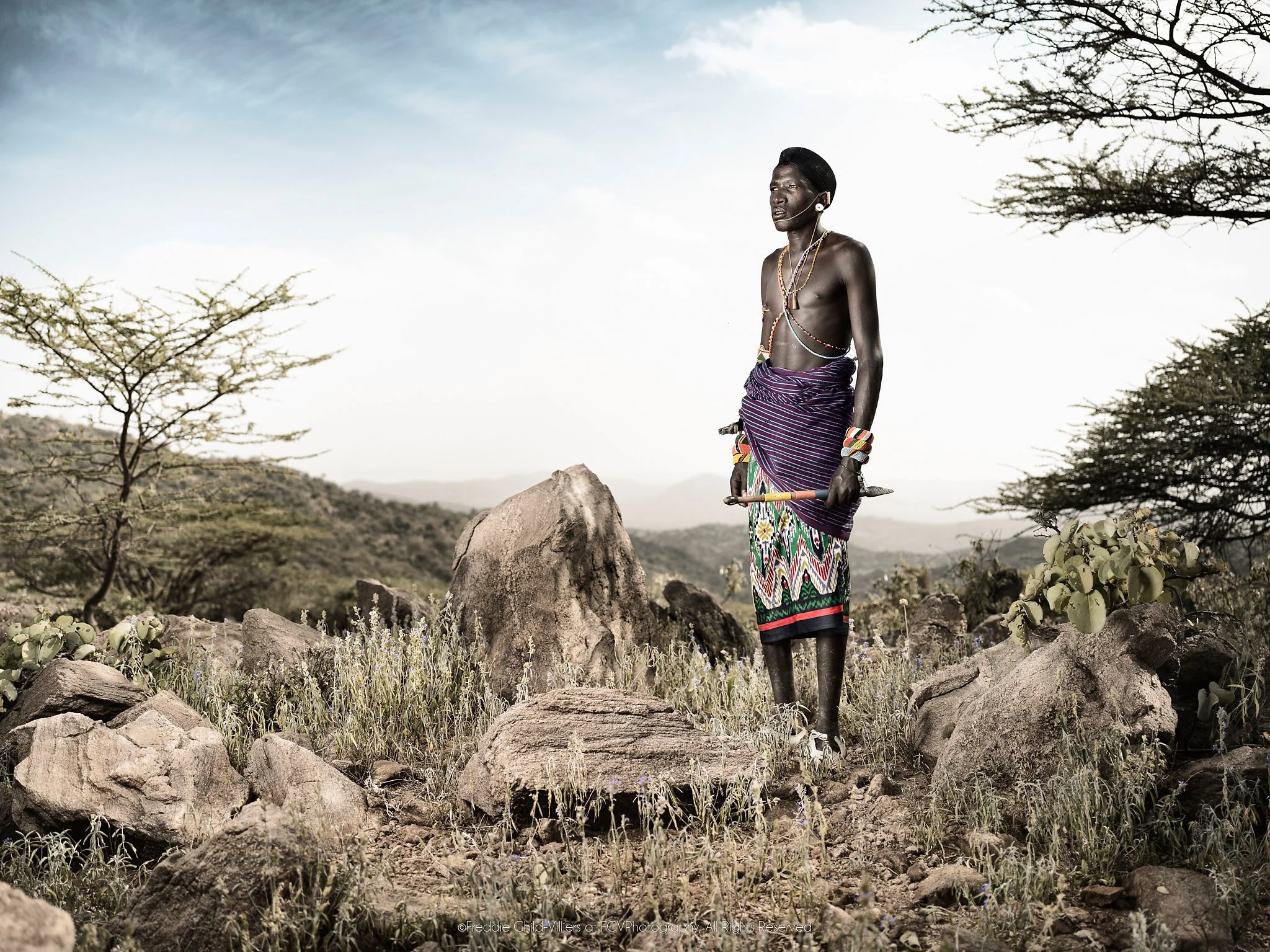Lkatinga, a Samburu man in purple and patterned clothing standing among rocks in the Matthews Mountains.