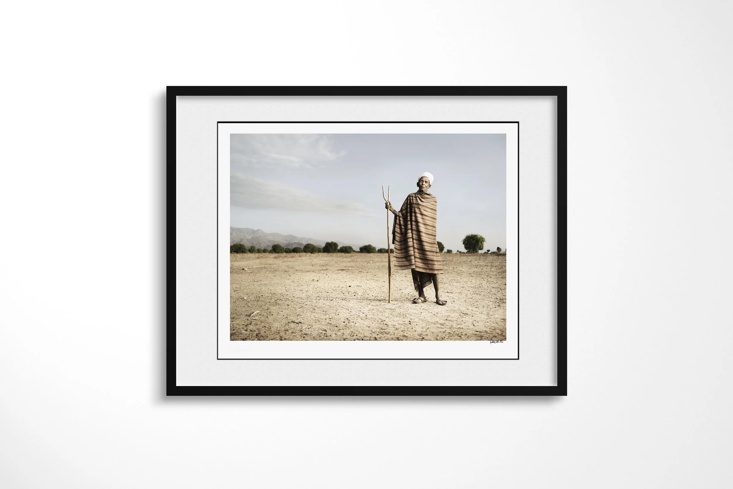 Framed presentation of Aregodo in a black frame and white mount, showing an older Arbore man in a striped wrap and white head cloth holding a forked staff on a dry plain.