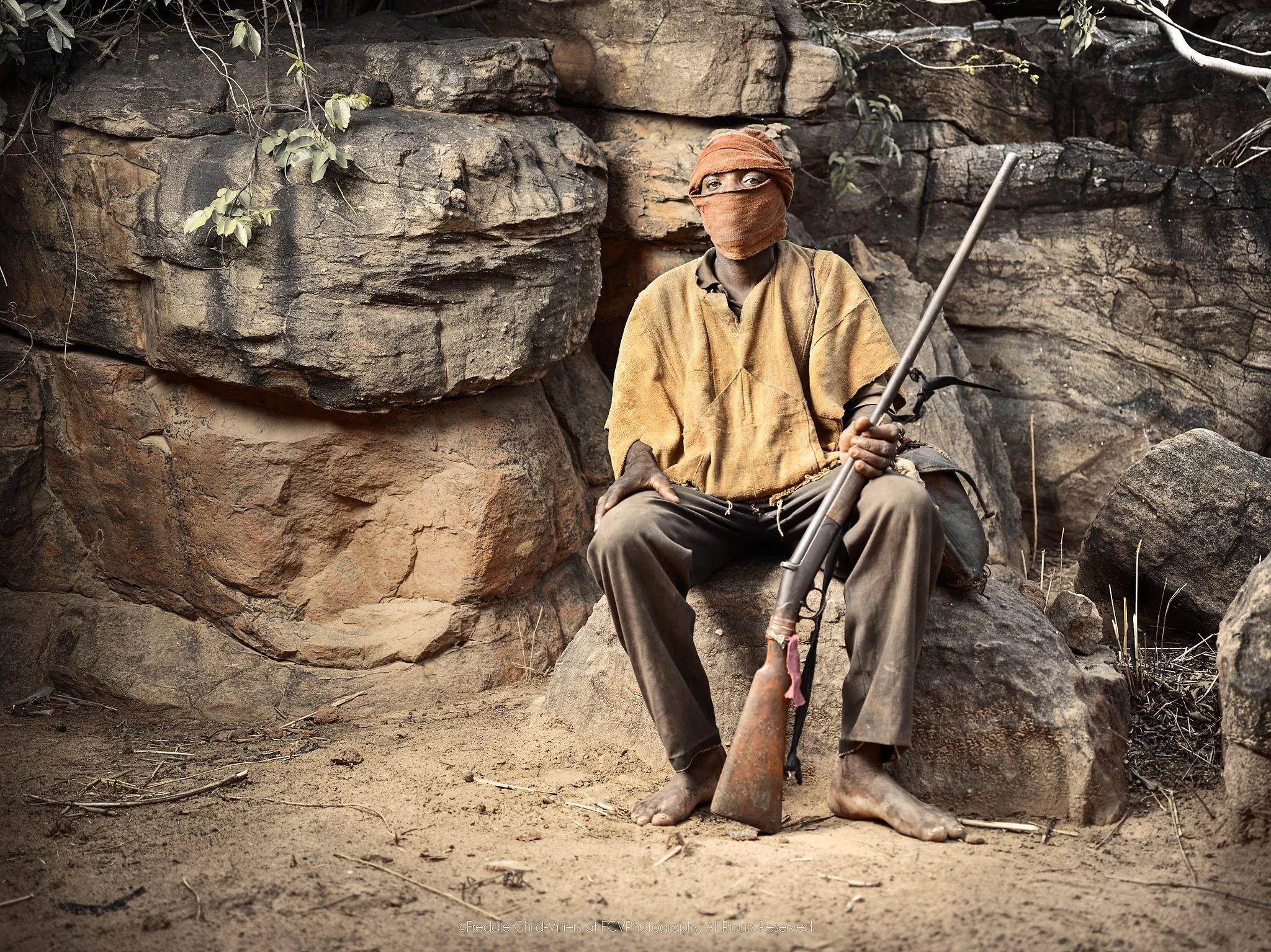 Duogolou seated barefoot against a rock face with his face wrapped and a rifle in hand.