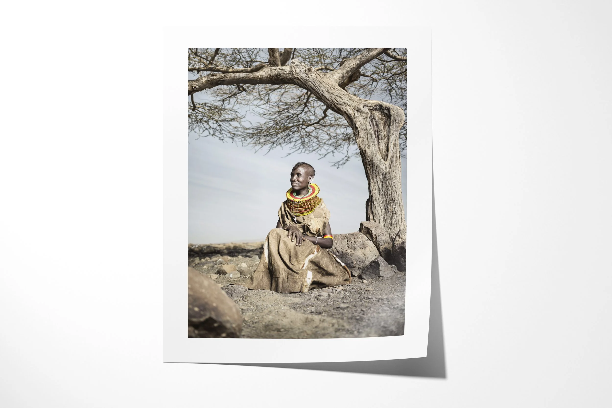 Fine art paper print of Lotilan, showing a Turkana woman in a hide dress seated beneath an acacia in rocky country.