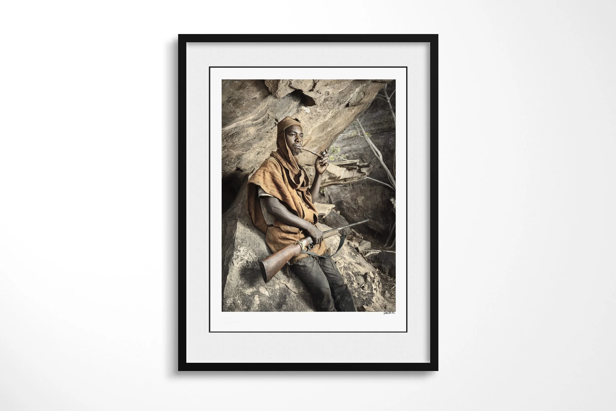 Framed presentation of Anbile in a black frame and white mount, showing Anbile seated in a rocky shelter smoking a long pipe with a rifle across his lap.