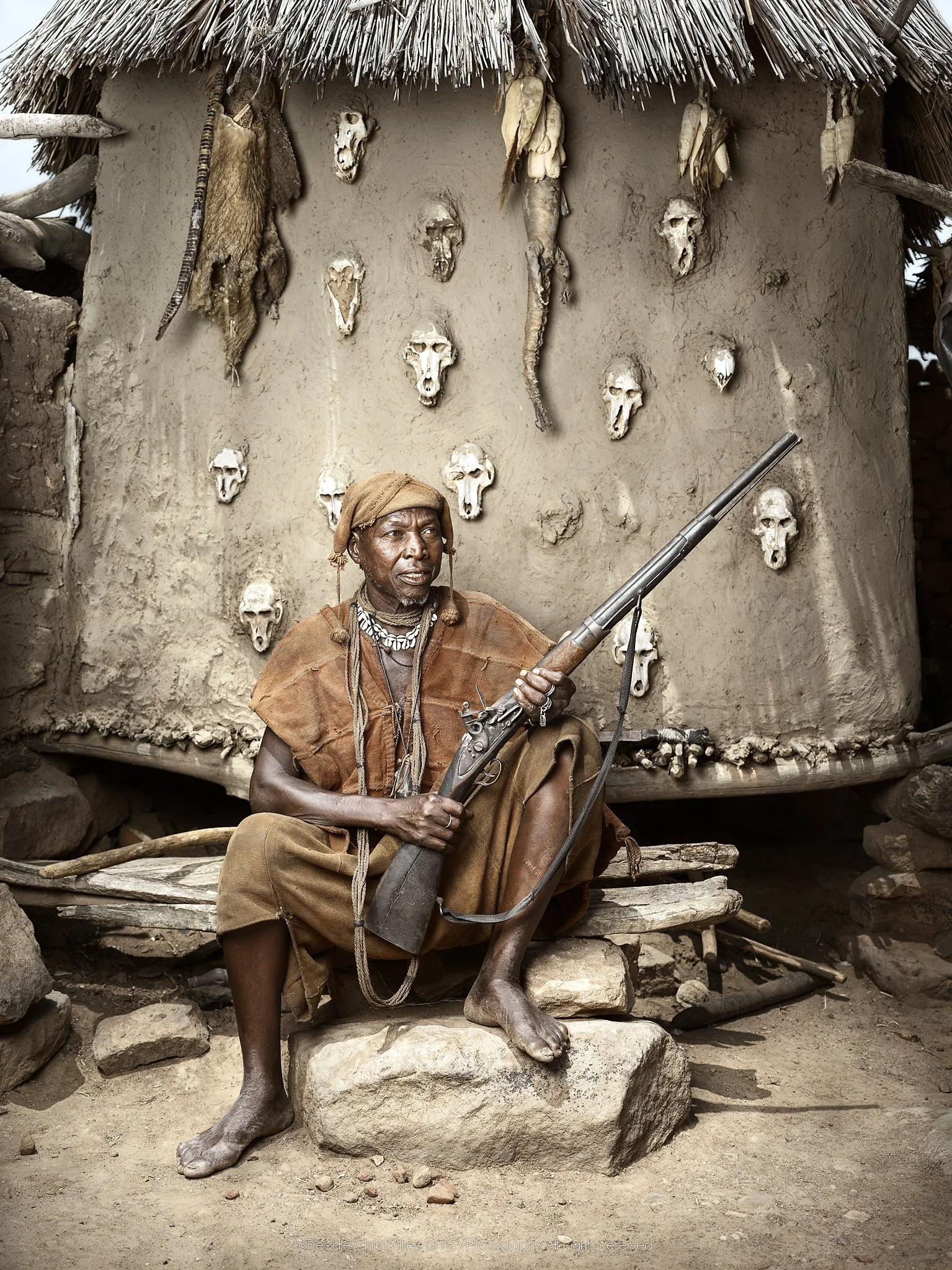 Lion seated with a flintlock rifle before an earthen wall set with animal skulls.