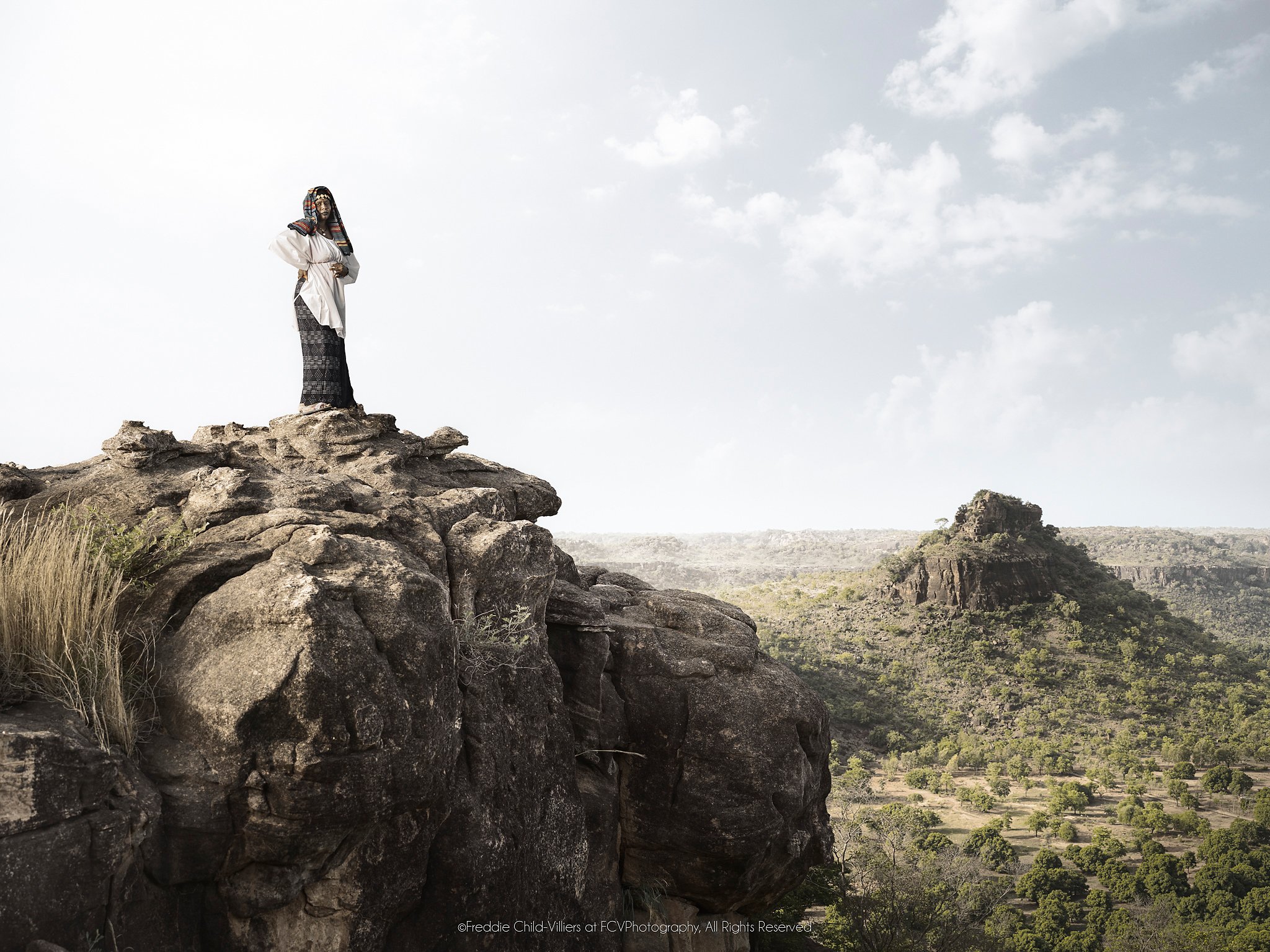 Fante standing on a high rocky ledge above a green valley and distant escarpment.