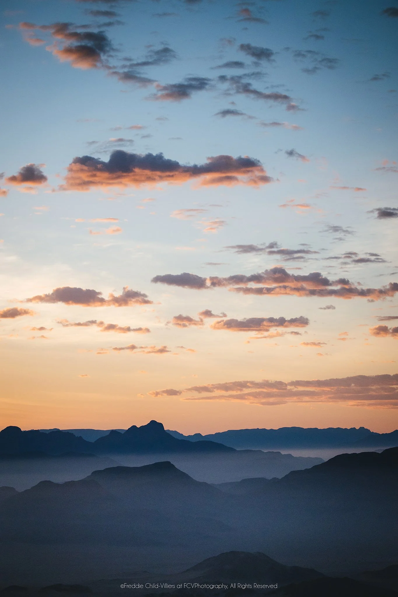 Layered dark desert ridges at Sossusvlei beneath peach and pale blue clouds at sunrise.