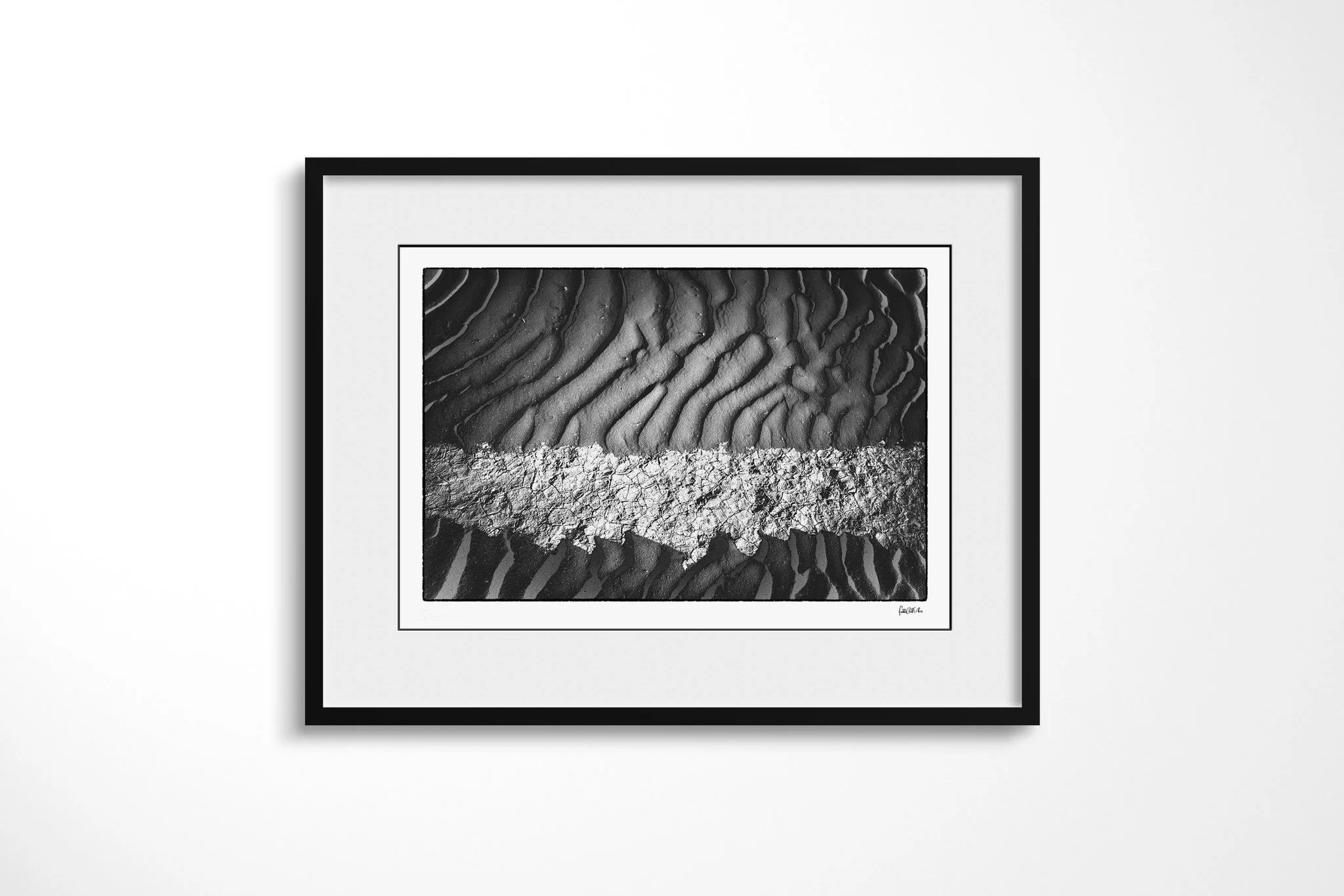 Khamsin II presented in a black frame and wide white mount, showing a pale cracked band crossing dark rippled sand in a high-contrast horizontal composition.