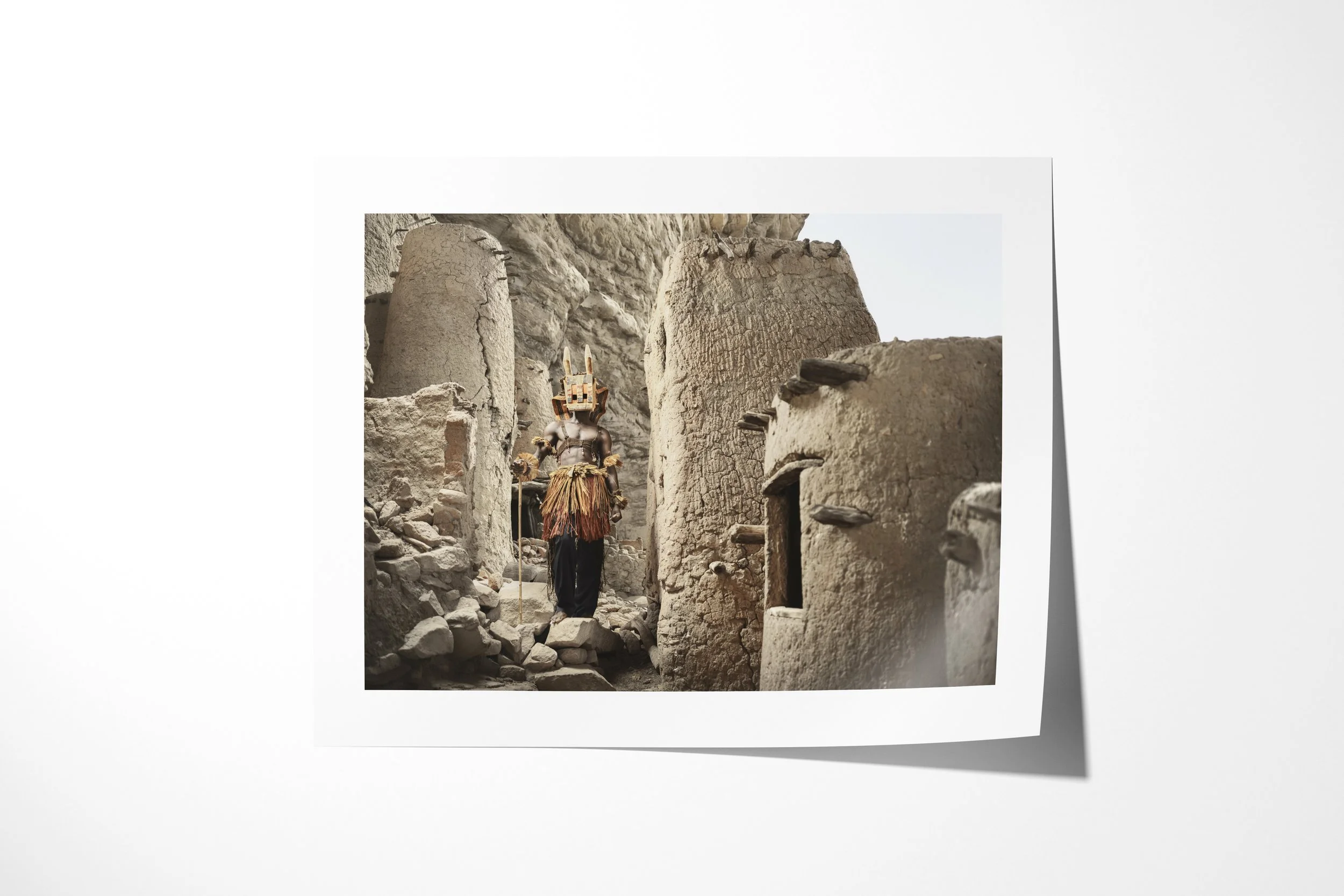Fine art paper print of Djomo (Mask), showing a Dogon Djomo mask moving through a narrow passage between earthen and stone buildings.