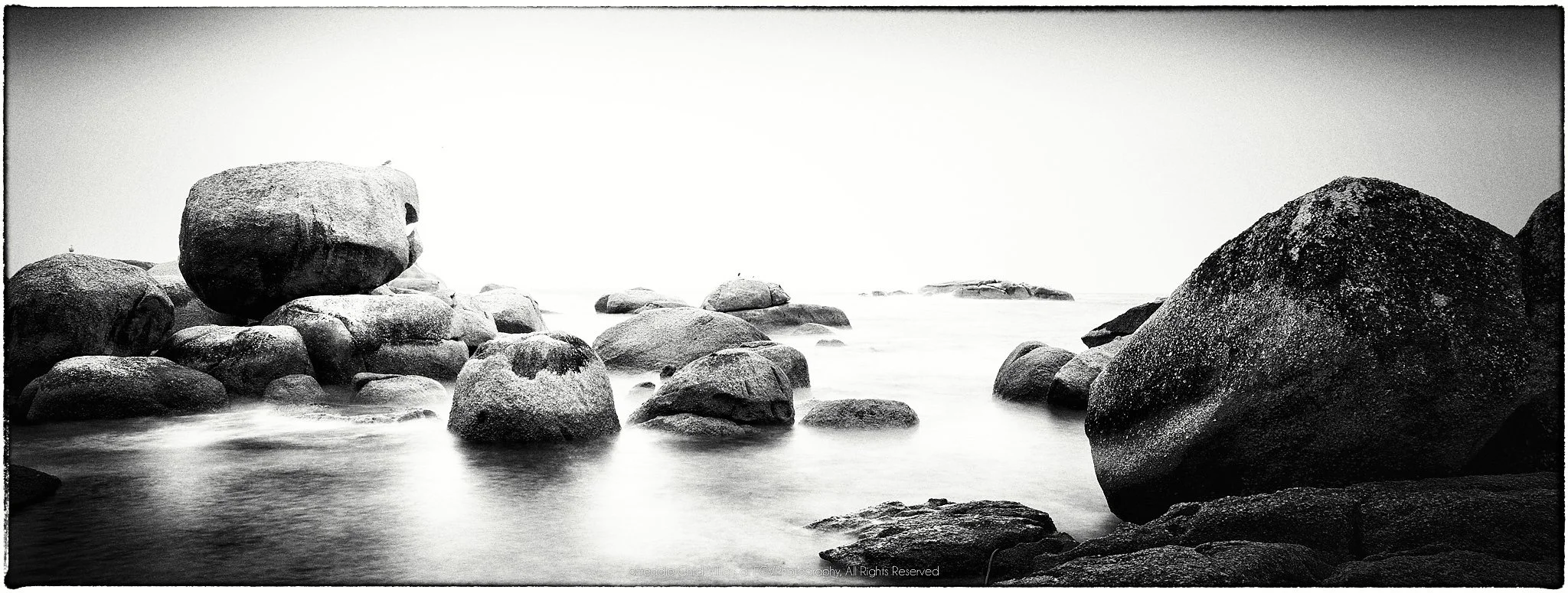 Wide black-and-white panorama of rounded coastal boulders emerging from smooth long-exposure seawater.