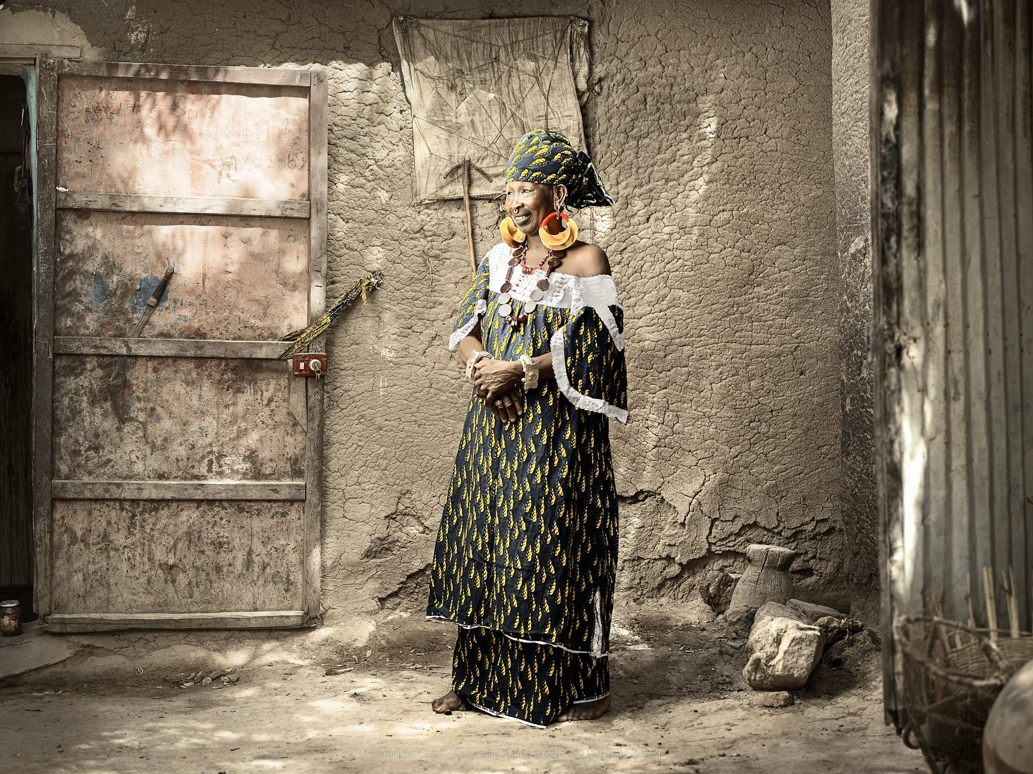 Thindel wearing large gold earrings and patterned clothing beside a weathered metal door.