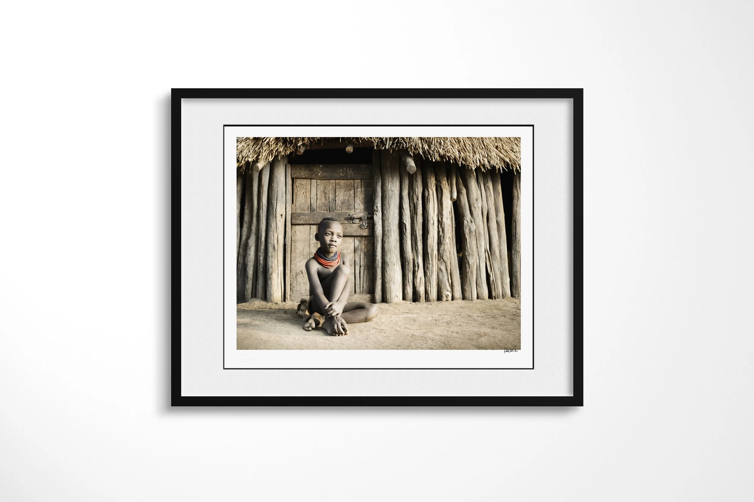 Framed presentation of Kayo in a black frame and white mount, showing a Kara child seated before the timber doorway of a thatched family house.