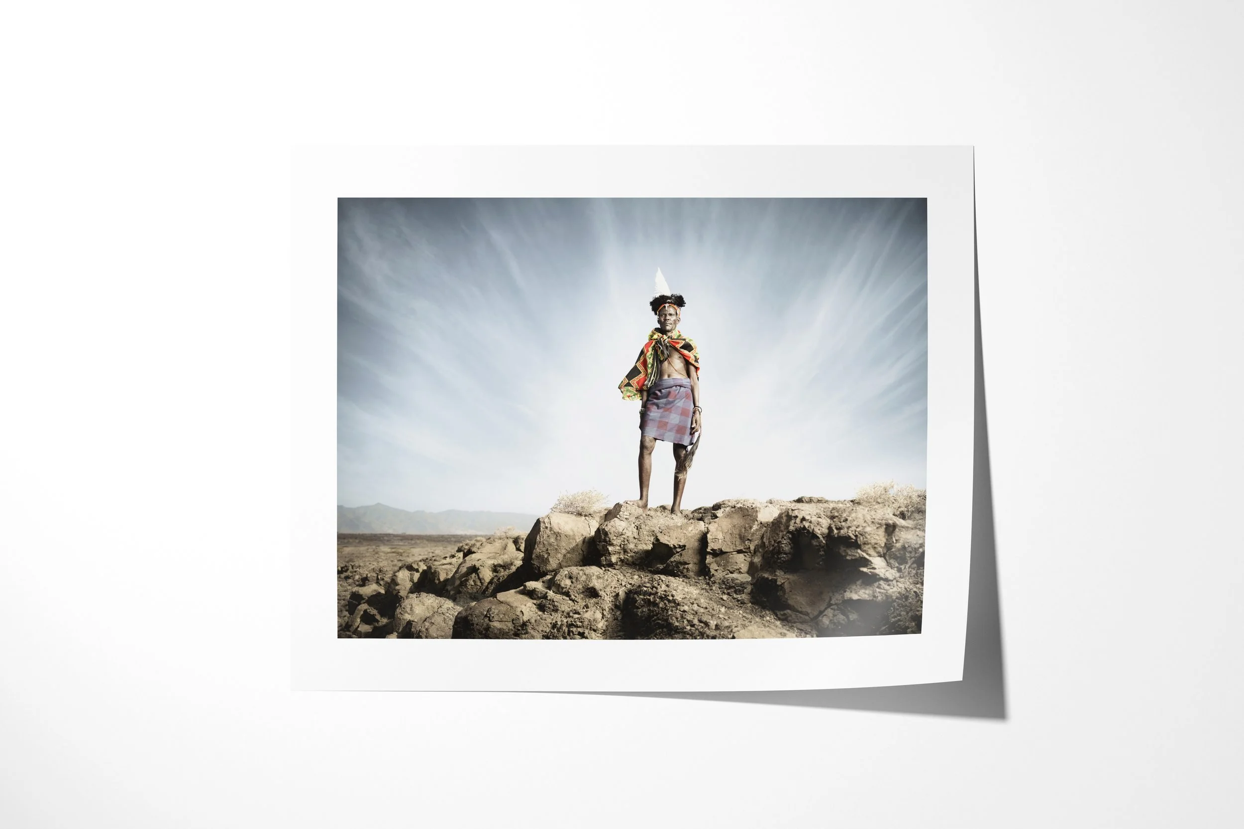 Fine art paper print of Ayapan, showing Ayapan standing on dark rocks in a feathered headdress beneath a streaked sky.