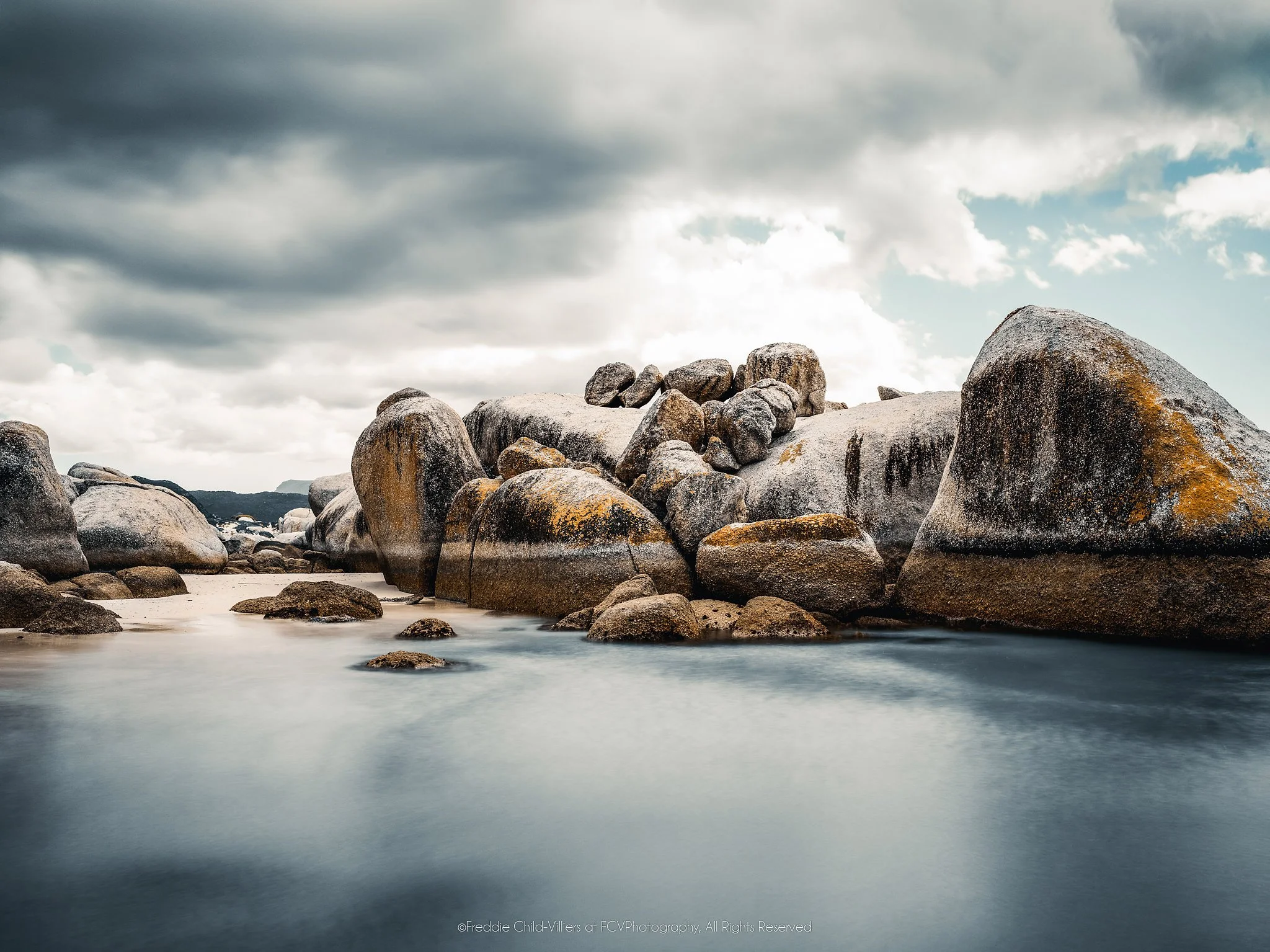 Rounded granite boulders around smooth blue-grey coastal water beneath heavy clouds, with patches of warm lichen.