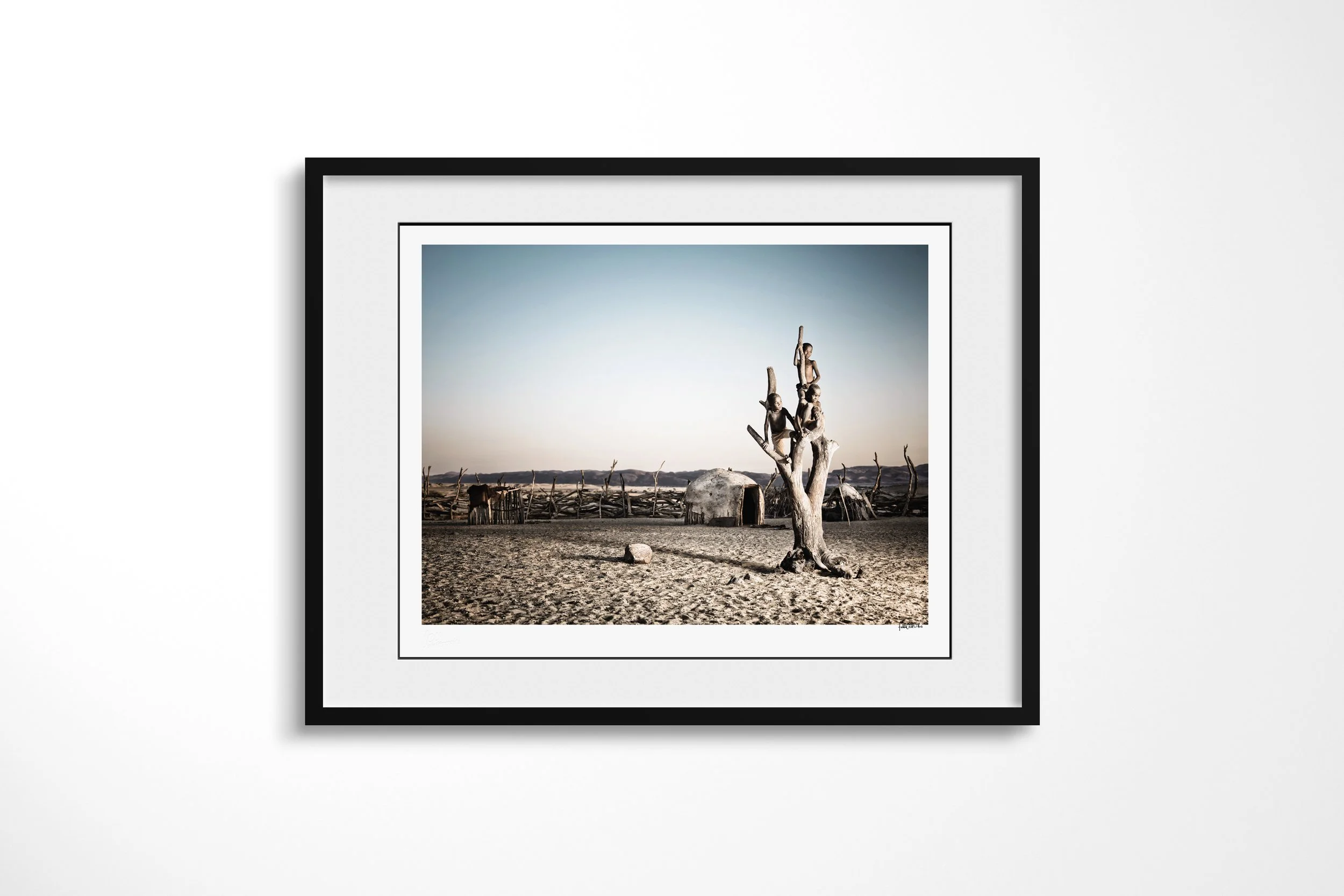 Framed presentation of The Tree in a black frame and white mount, showing three Himba boys climbing and sitting in the bare branches of a tree inside a homestead.