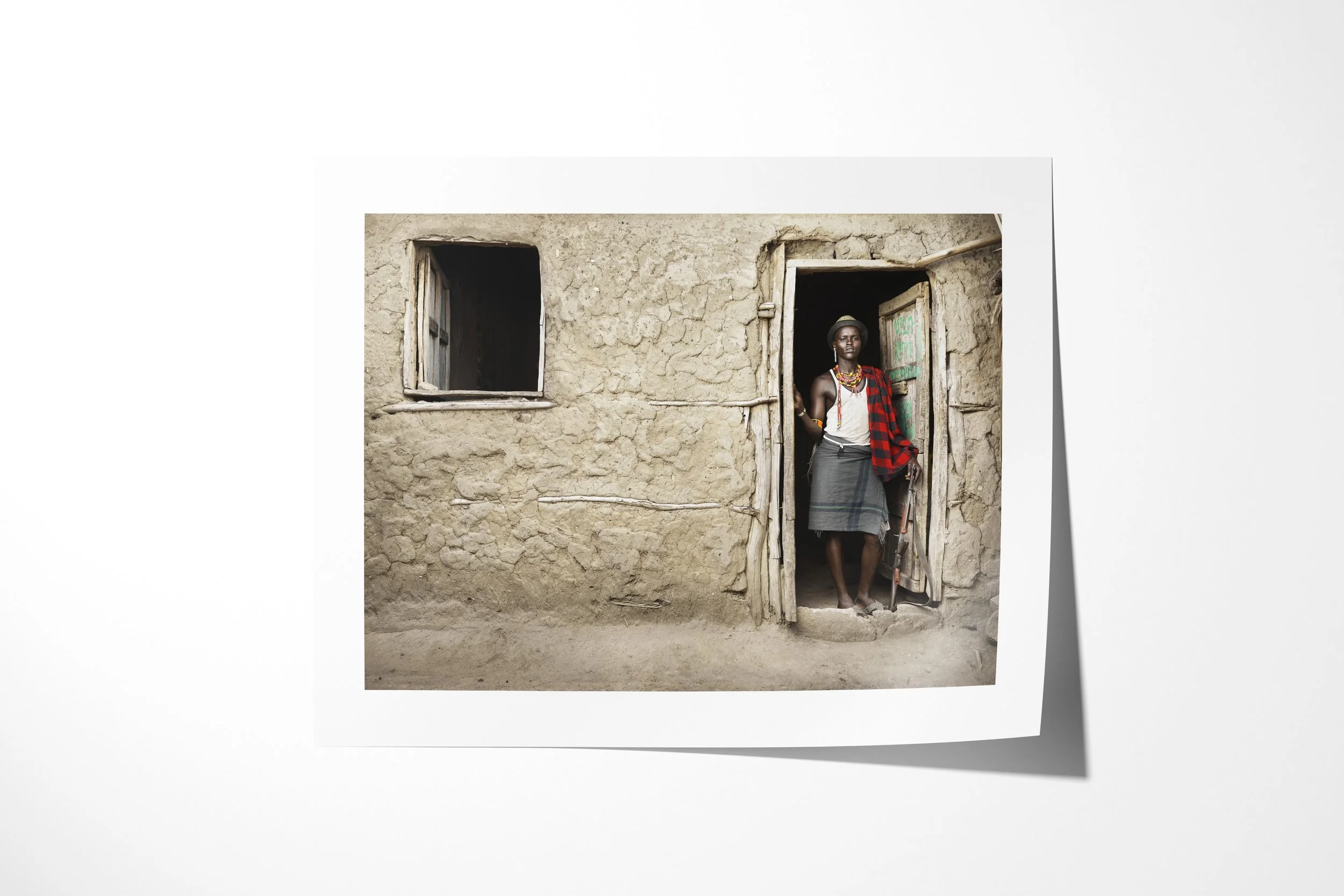 Fine art paper print of Arboro, showing an Arbore man standing in the doorway of a mud-walled house wearing beadwork and a checked cloth.