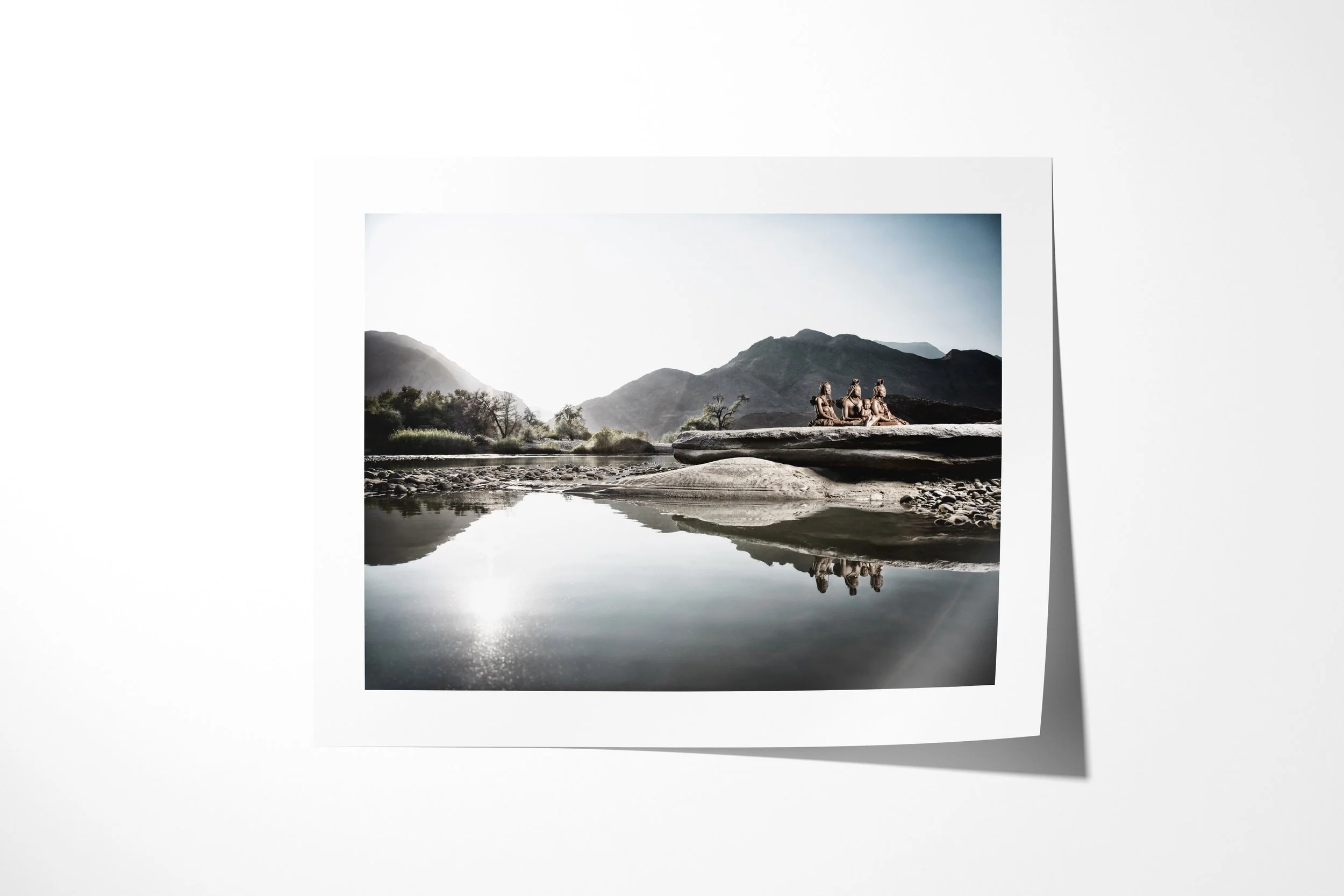 Fine art paper print of Mbendura (left), Tjikuria and child (middle), Mutume (right), showing a Himba family gathered on a broad rock beside the Kunene River with their reflections in the water.