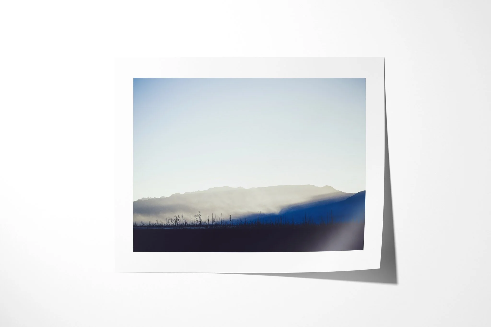 Fine art paper presentation of Drought, depicting bare blackened trees below a mountain ridge, with pale mist lit above a deep blue shadow.