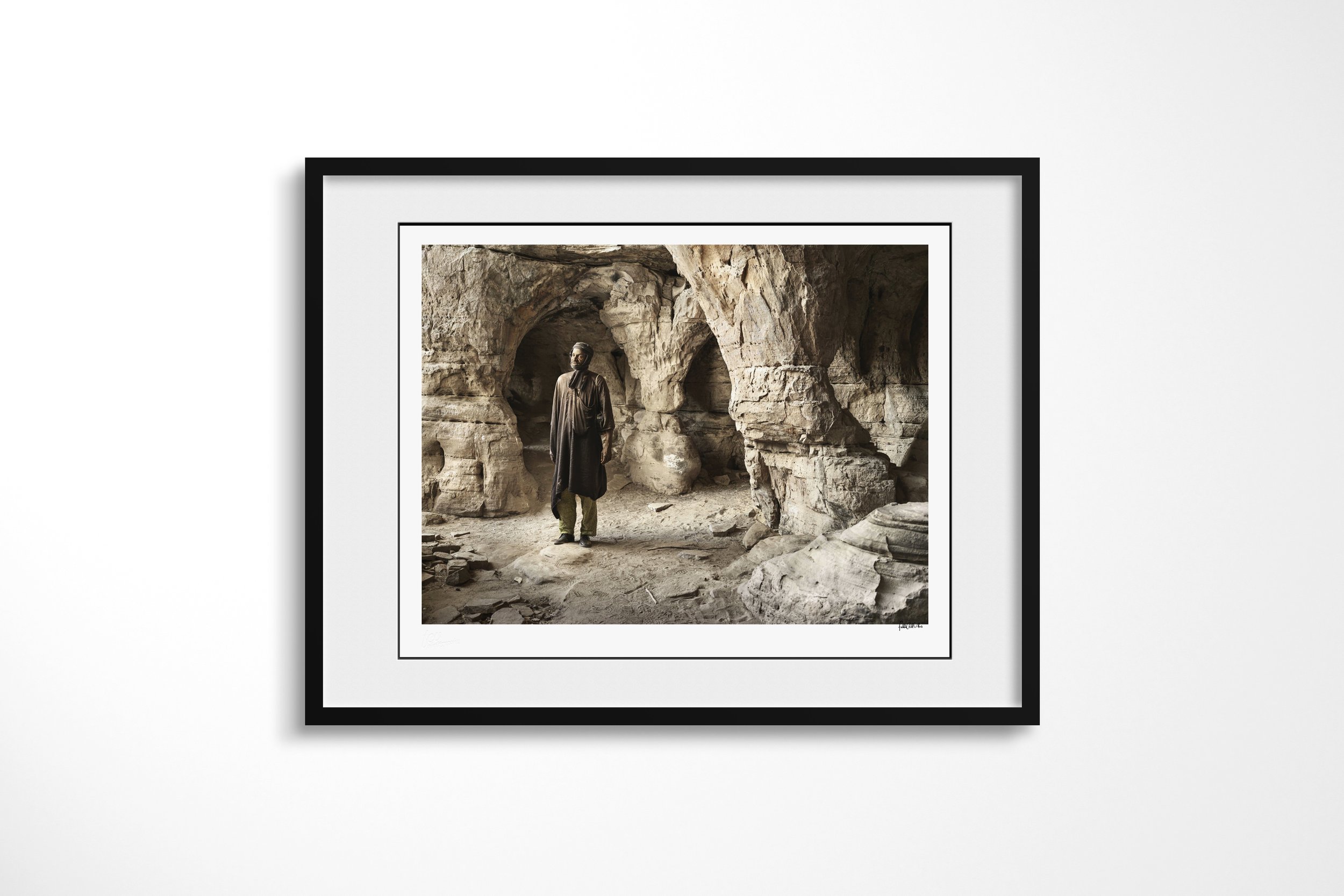 Framed presentation of Alou in a black frame and white mount, showing Alou standing within the carved arches and columns of a sandstone cave.