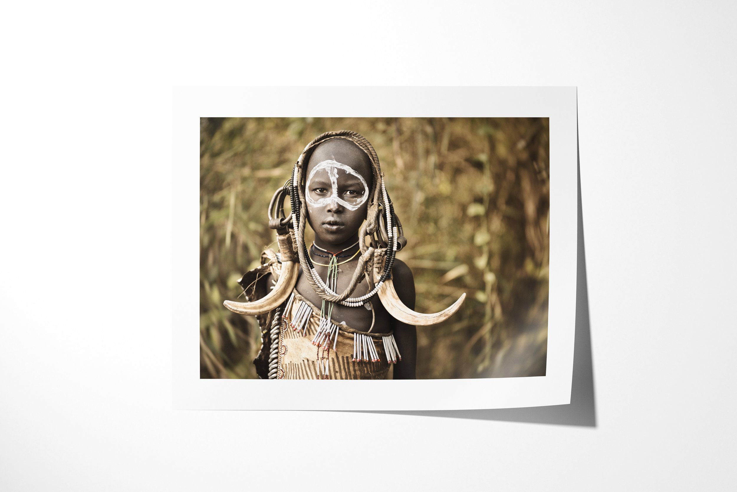 Fine art paper print of Ngateri, showing a young Mursi girl with white facial paint, beadwork and paired tusk ornaments against woodland.