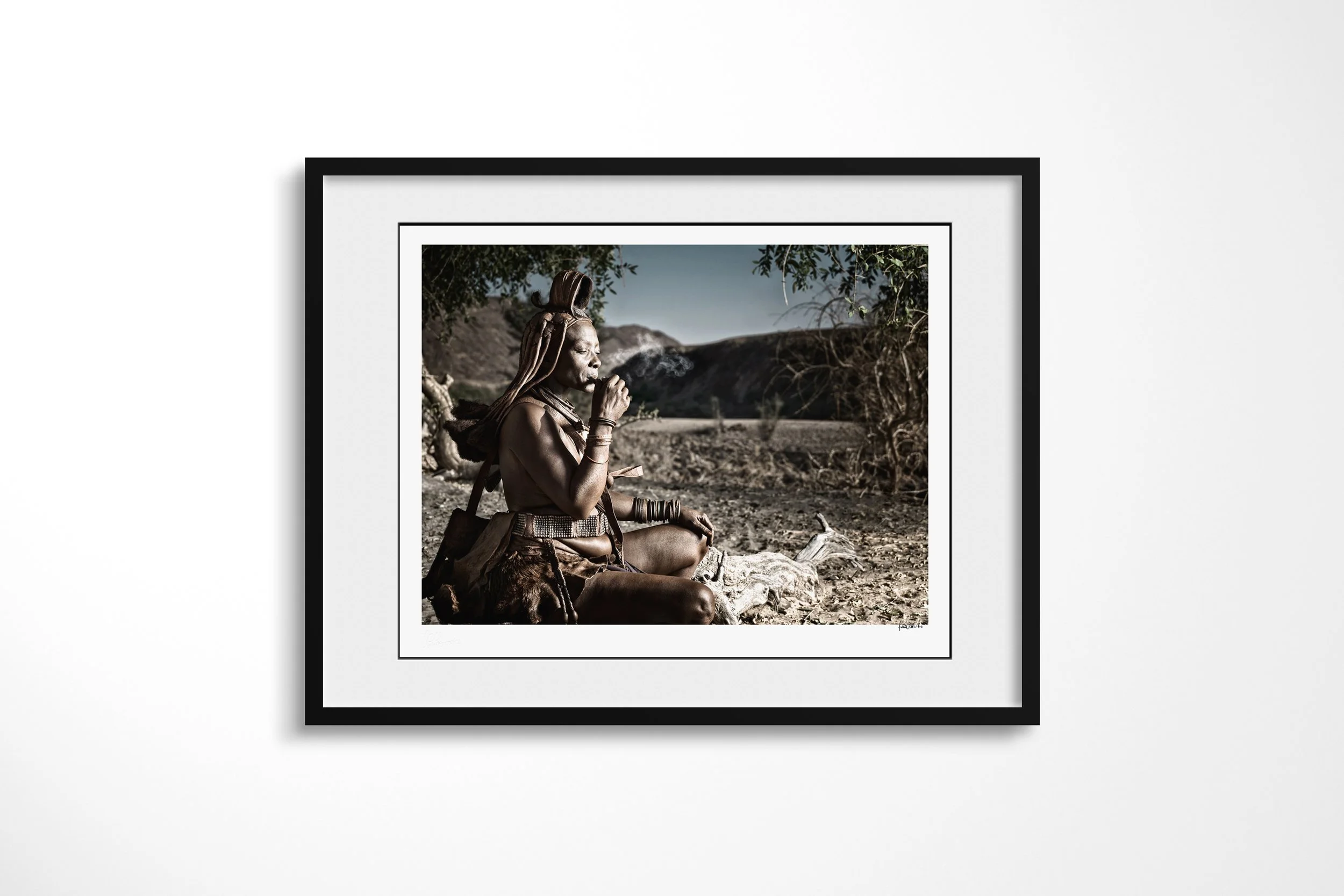 Framed presentation of Ngorera in a black frame and white mount, showing Ngorera seated beneath a tree smoking a small pipe in the dry Kunene landscape.