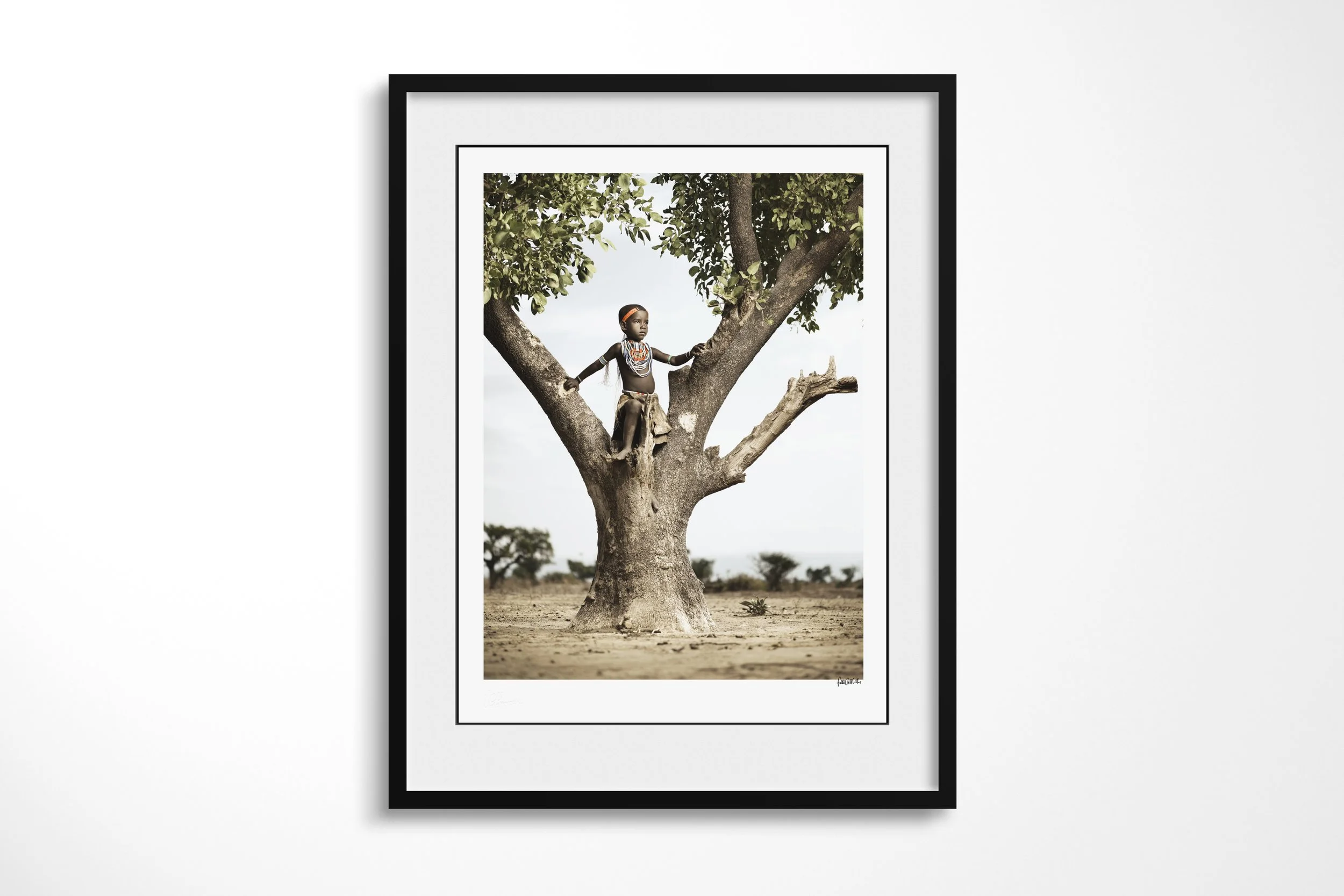 Framed presentation of Kenina in a black frame and white mount, showing a young Arbore girl standing in the fork of a large tree above dry ground.