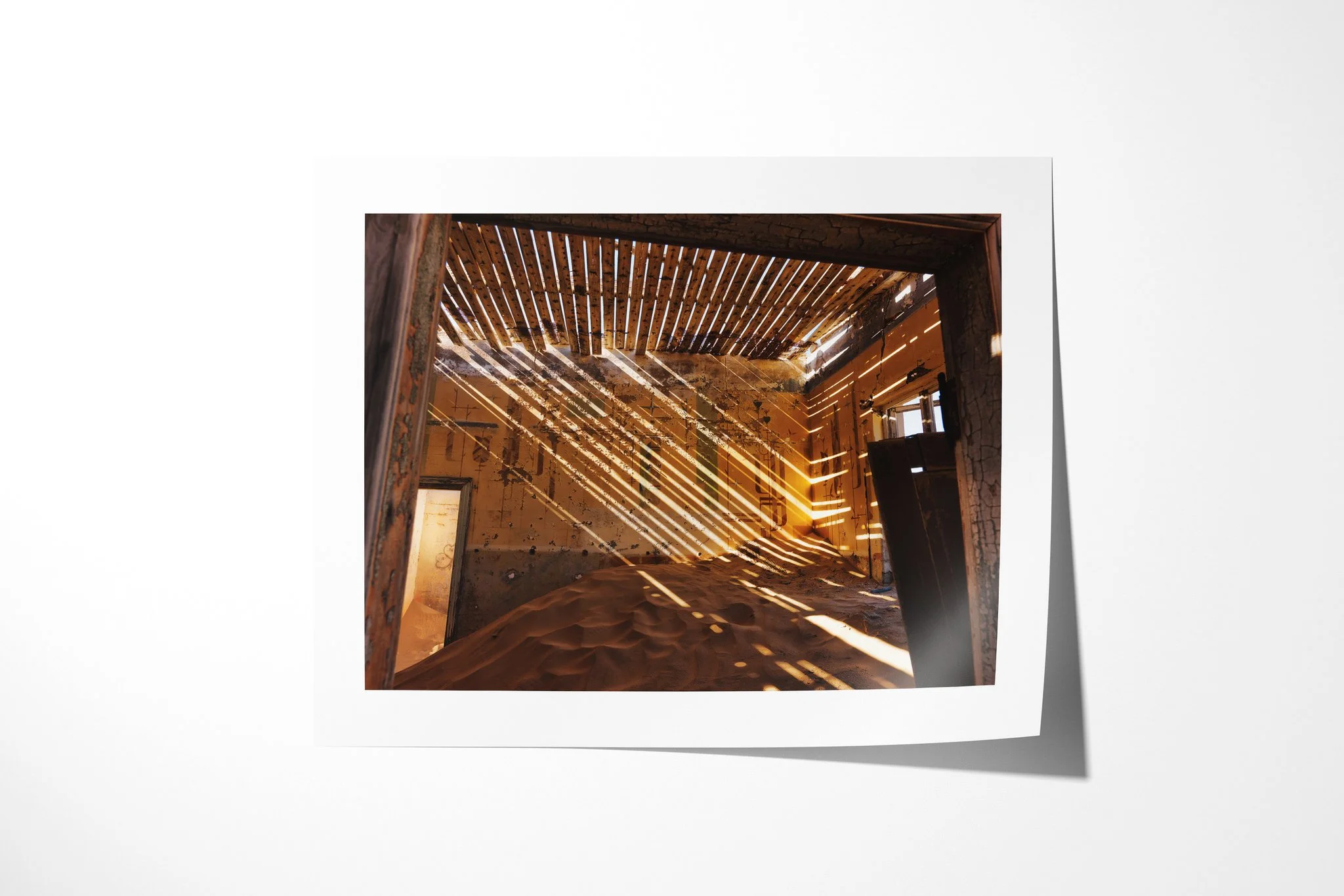 The Ministers House presented as an unframed fine art paper print, depicting a sand-filled ochre room crossed by diagonal sunlight from a damaged slatted roof, with peeling walls and an open doorway.