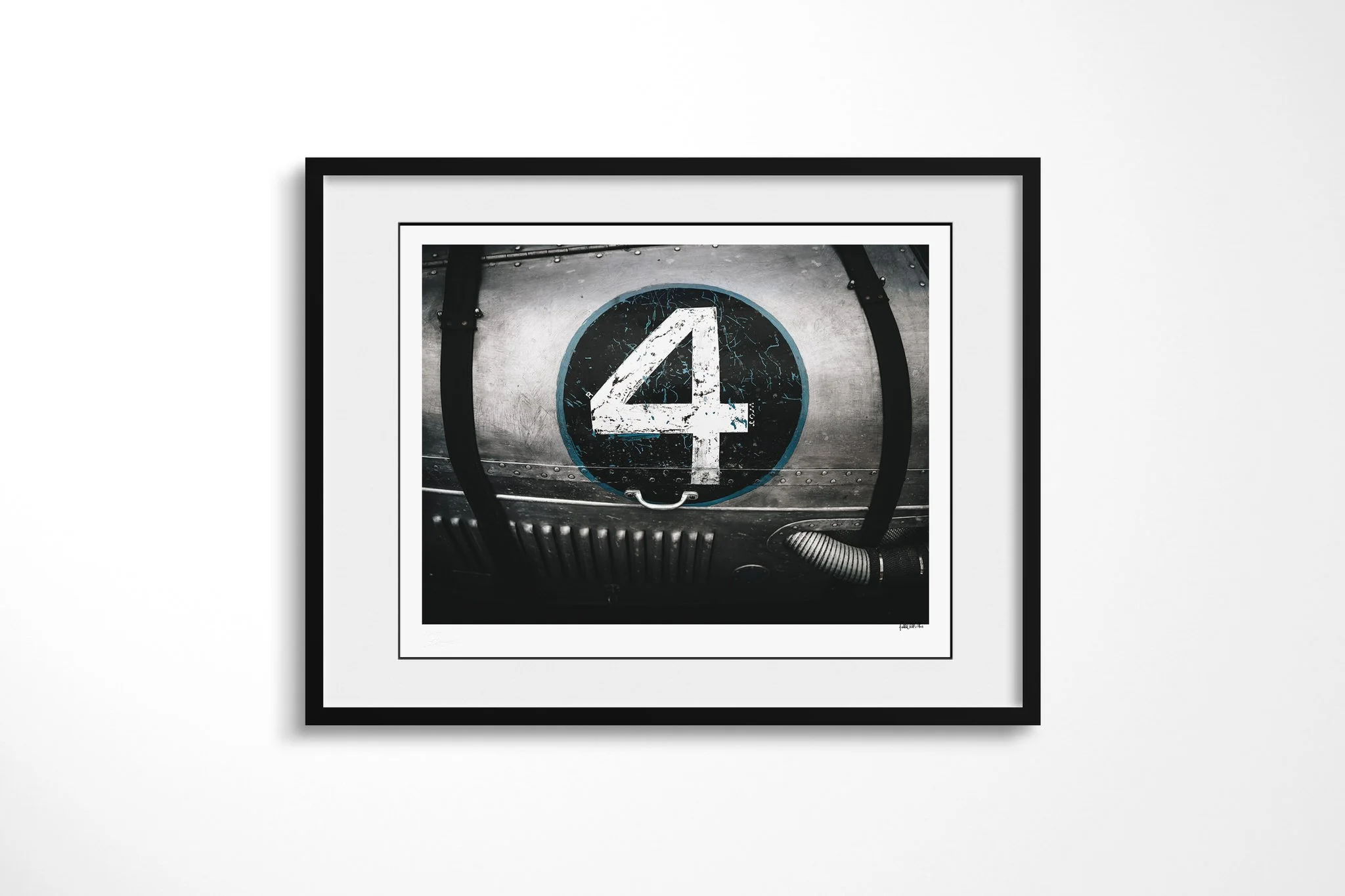 No.4 presented in a black frame with a white mount, depicting weathered white numeral four on a black circular plate set into riveted metal with straps and ribbed tubing.
