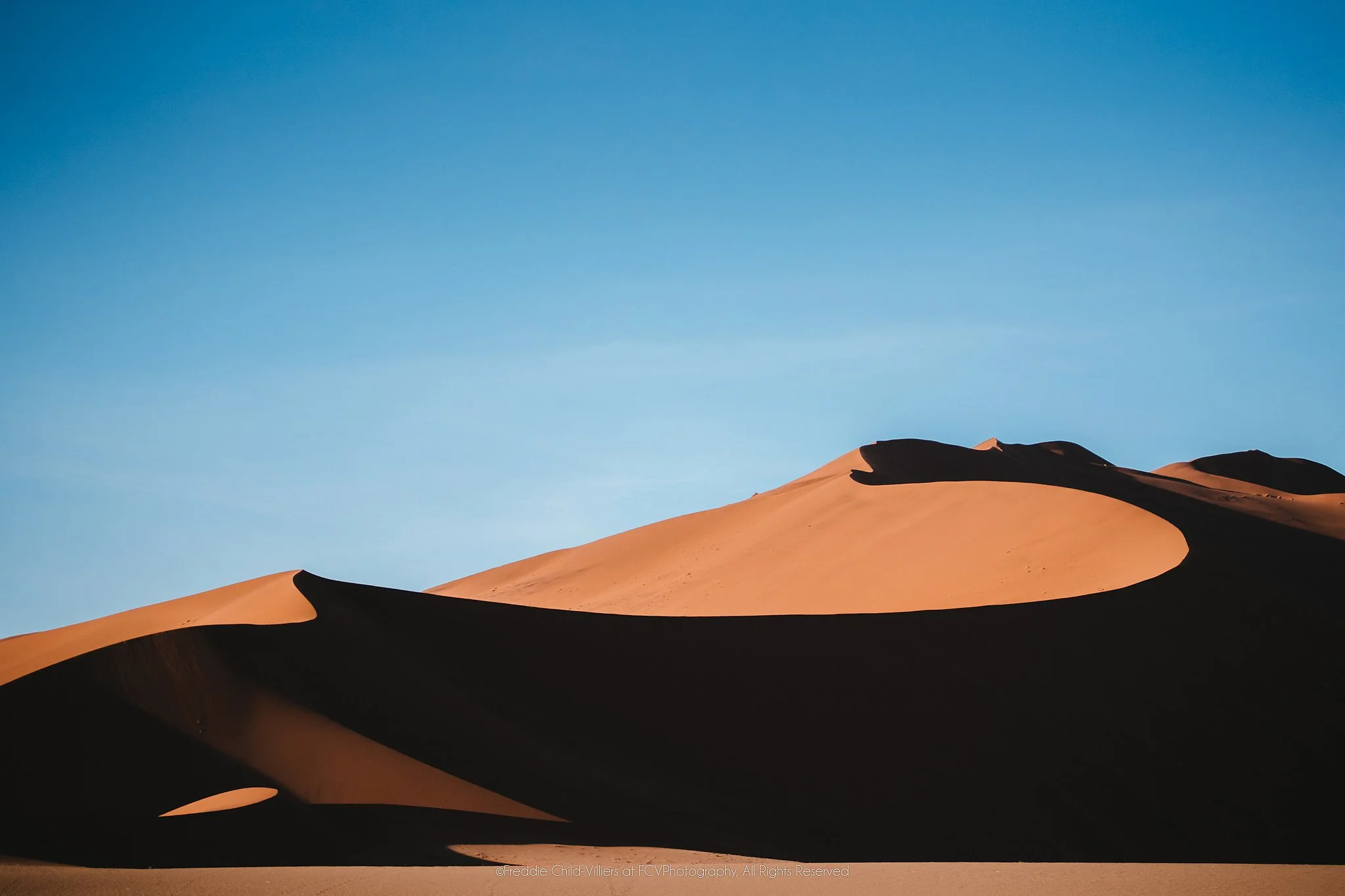 Sunlit orange desert dunes divided by deep sinuous shadows beneath a clear blue sky.