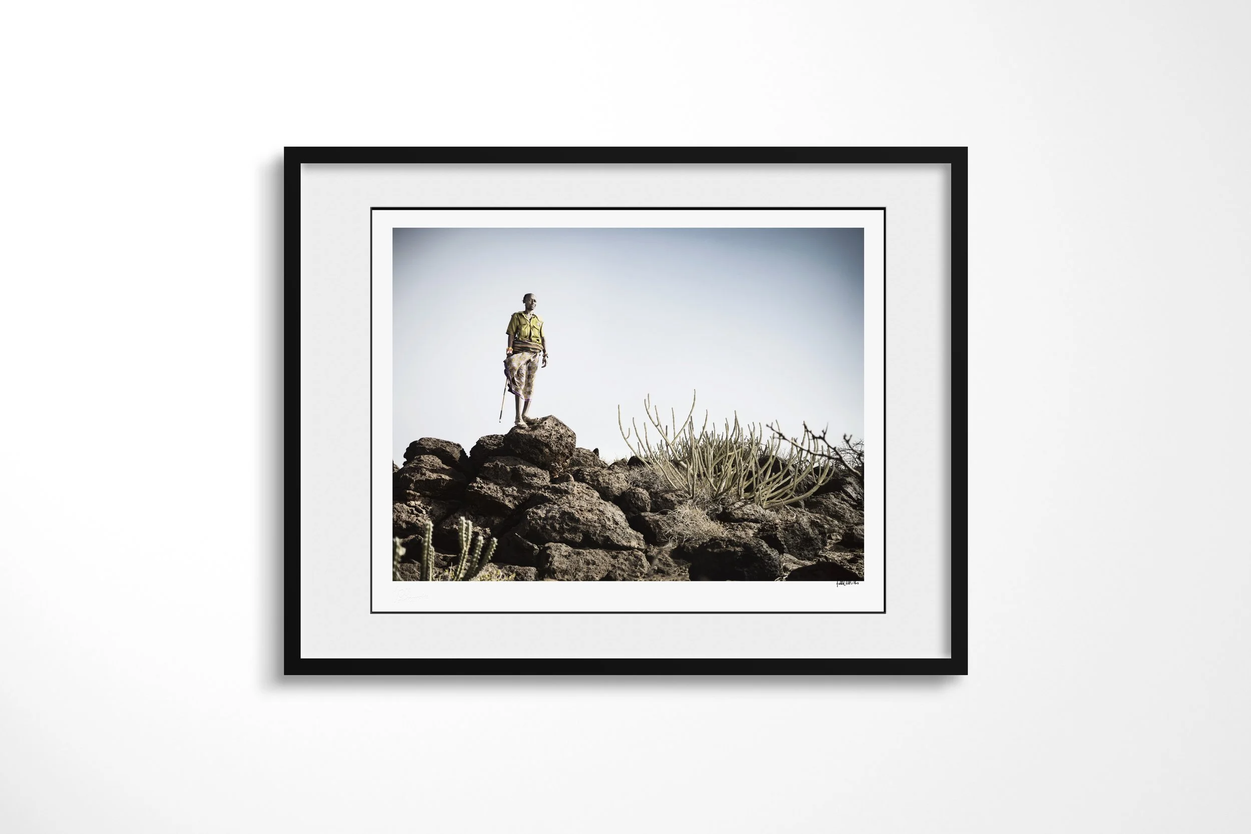 Framed presentation of Lpushe - “The Shepherd” in a black frame and white mount, showing a Rendille herder holding a staff on black rocks beside a desert succulent.
