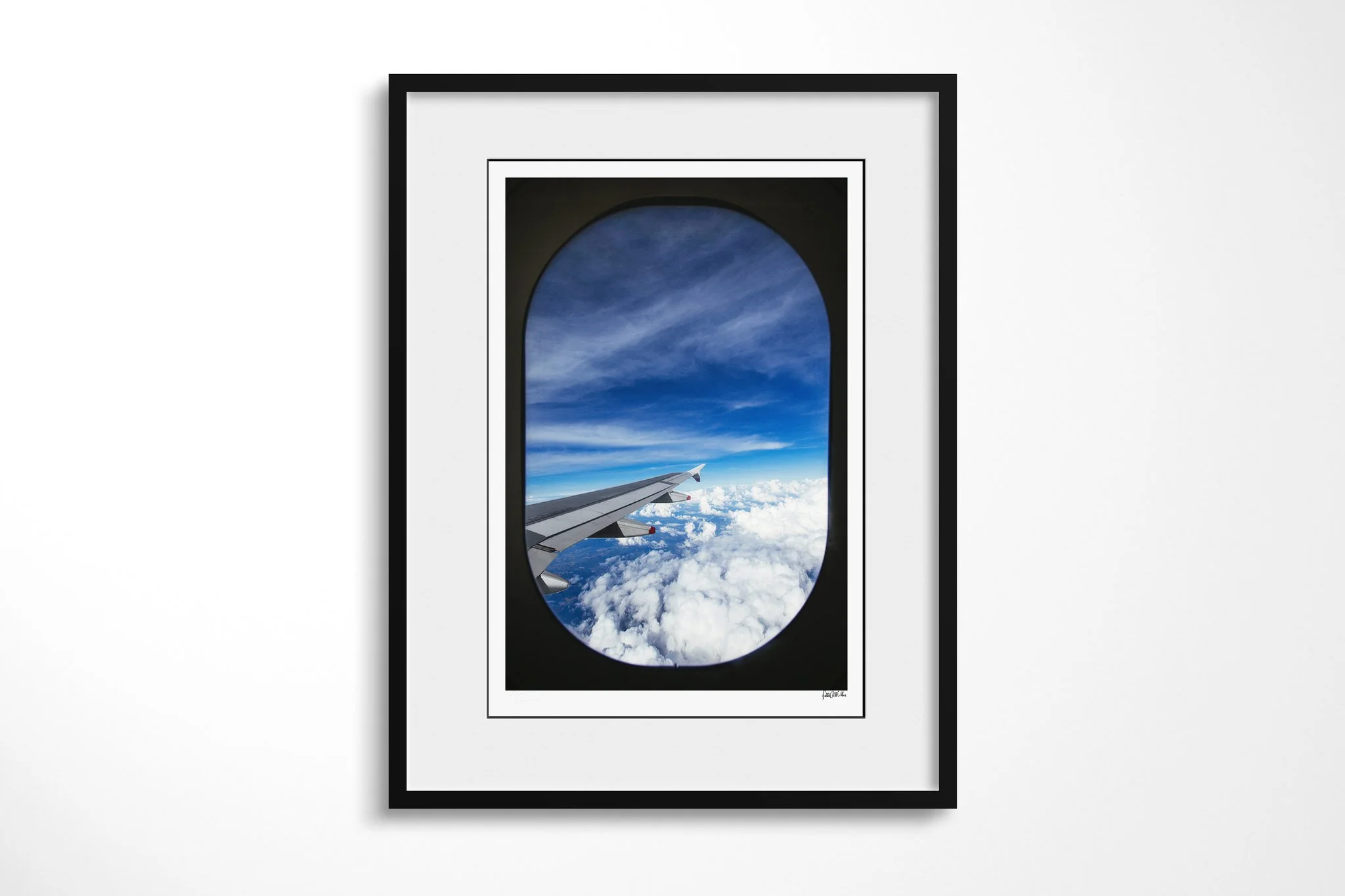 Black-framed fine art print of Room With A View with a white mount, showing an aircraft wing above bright cloud tops and deep blue sky, framed by the dark oval of a passenger window.