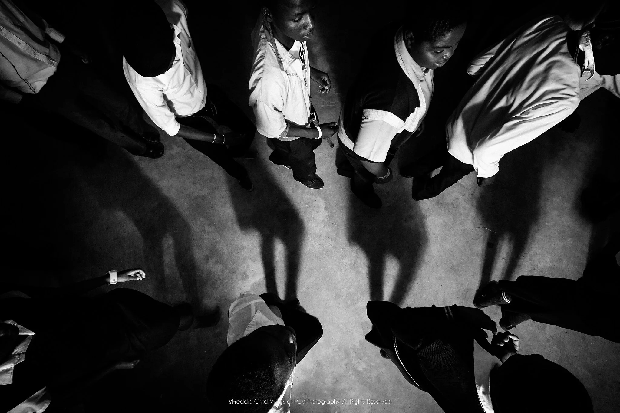 Group of people seen from directly overhead, standing in a loose circle with long shadows across a concrete floor.