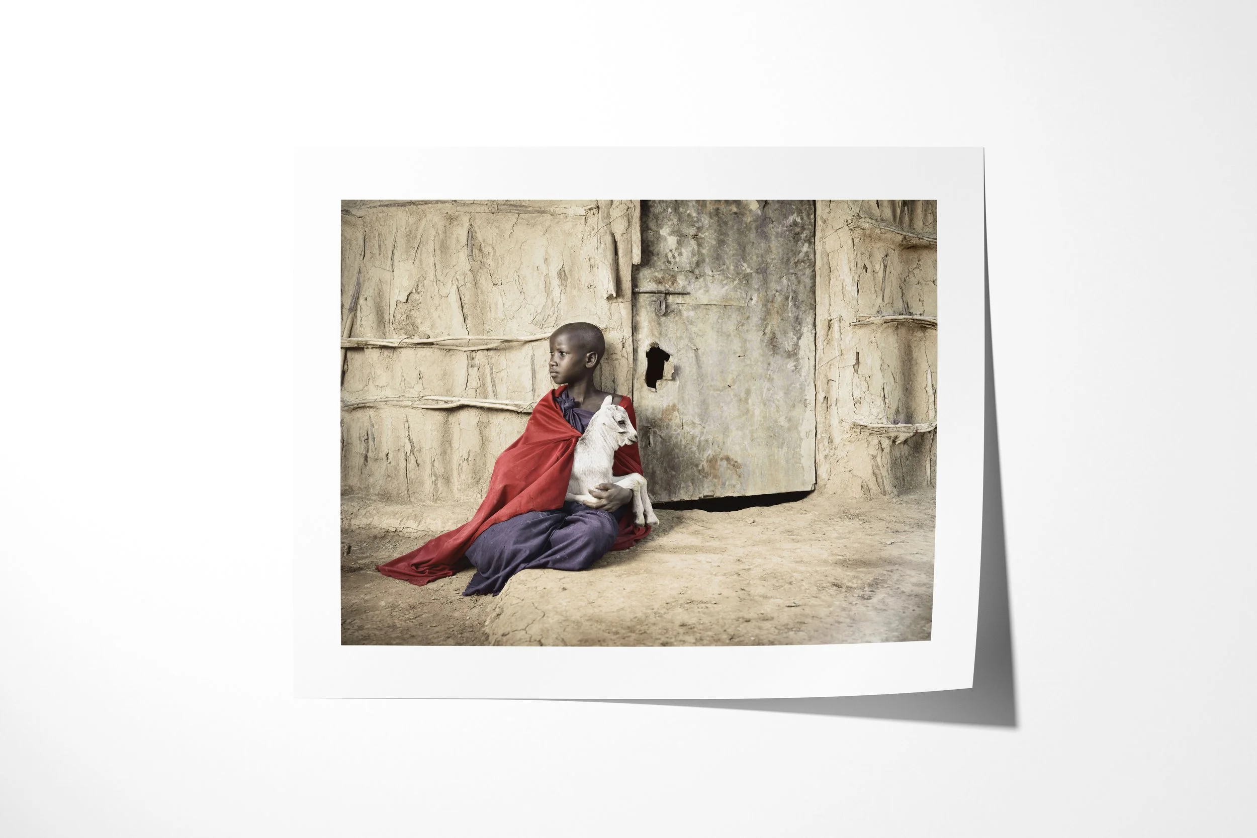 Fine art paper print of Nawasa, showing a Maasai girl wrapped in red cloth holding a young white goat outside an earthen house.