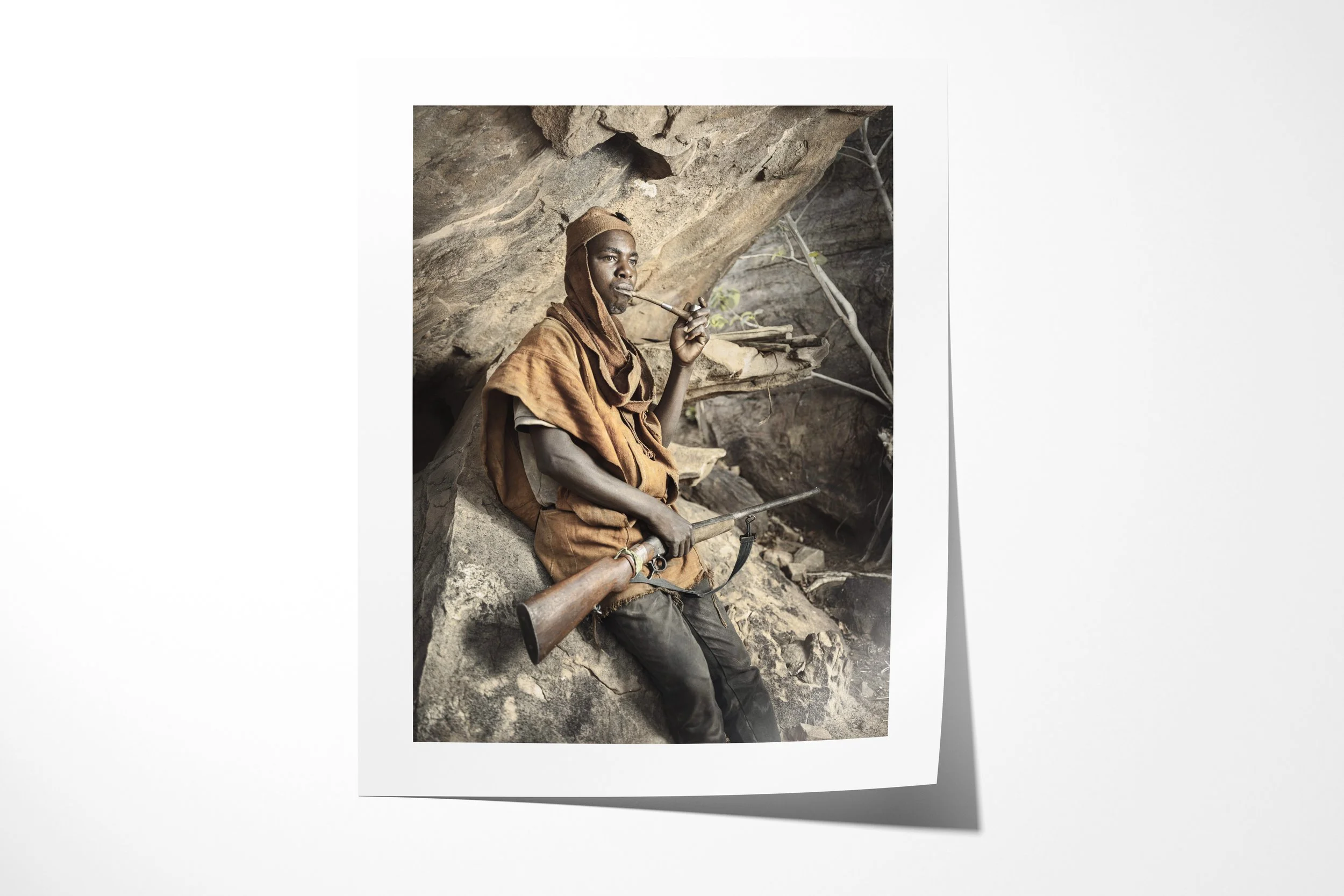 Fine art paper print of Anbile, showing Anbile seated in a rocky shelter smoking a long pipe with a rifle across his lap.