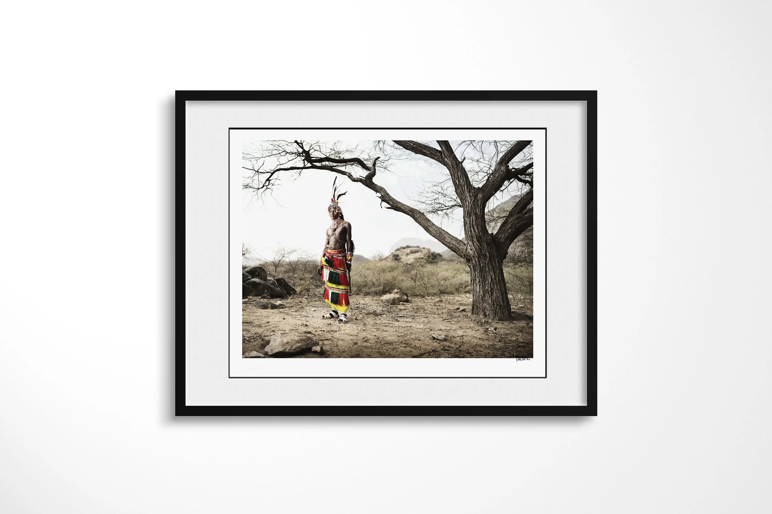 Framed presentation of Lebitili in a black frame and white mount, showing a Samburu warrior in colourful beadwork standing beneath a leafless tree in rocky country.