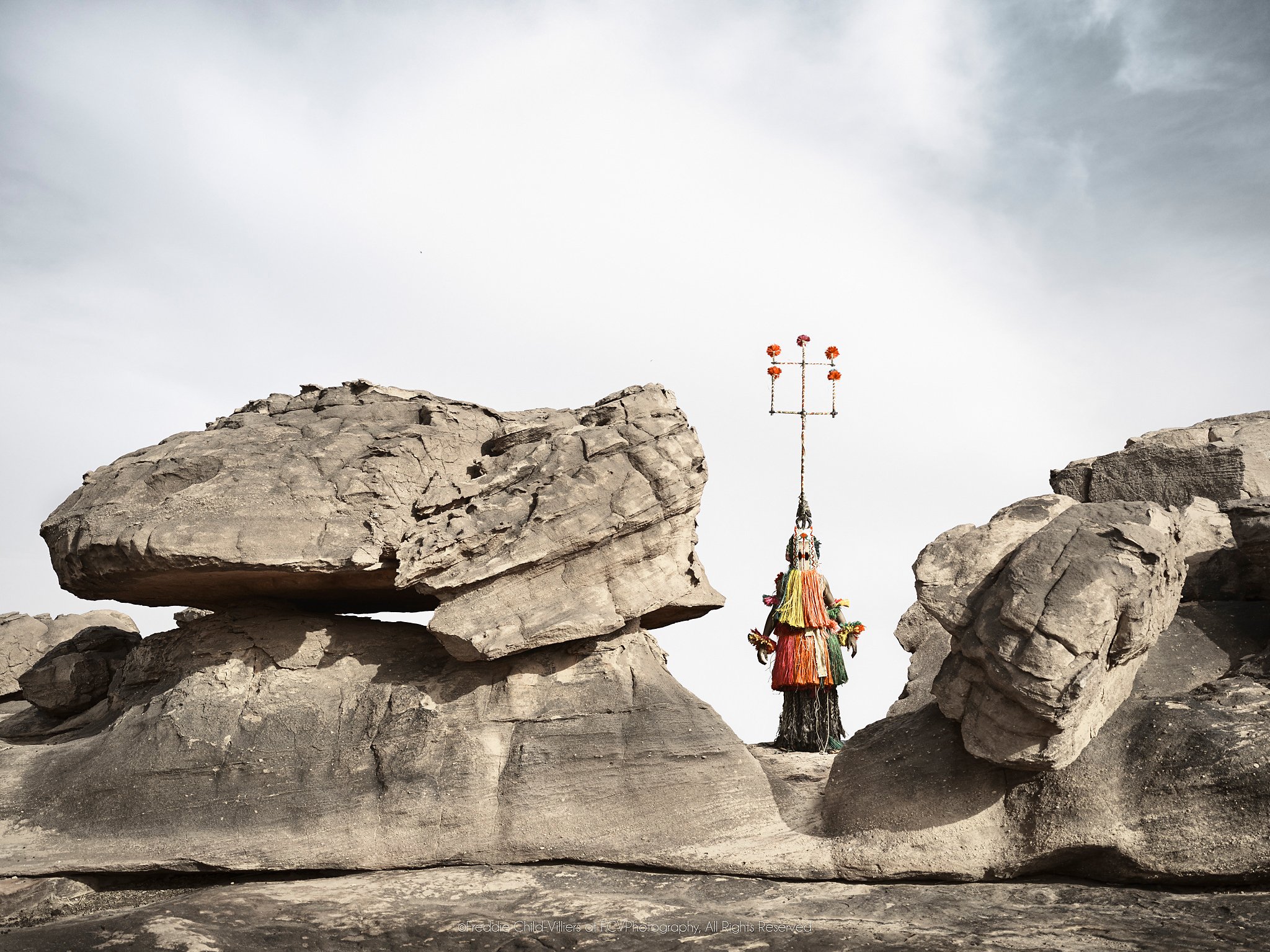 Karamagra (Mask), a tall Dogon Karamagra mask standing between massive boulders.
