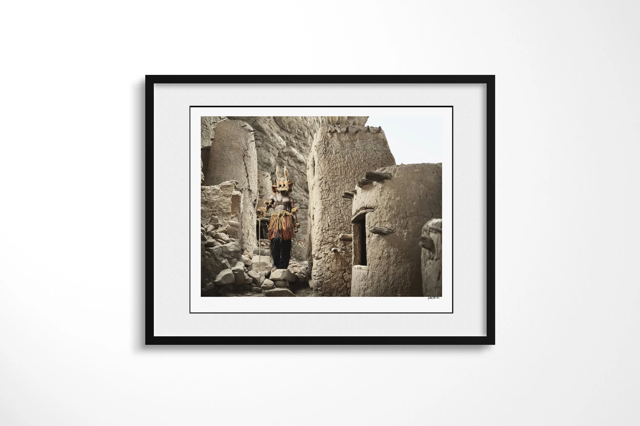 Framed presentation of Djomo (Mask) in a black frame and white mount, showing a Dogon Djomo mask moving through a narrow passage between earthen and stone buildings.