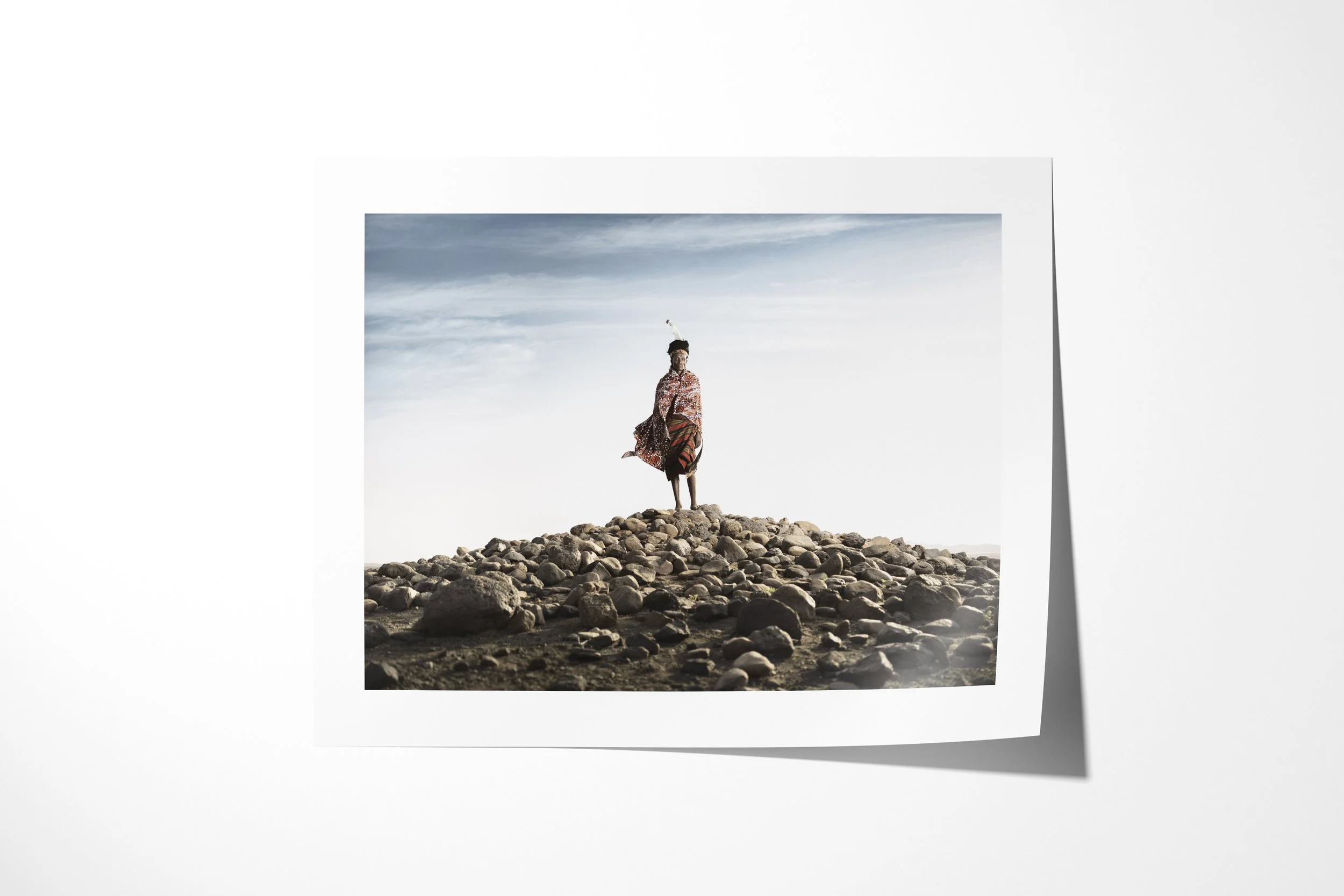 Fine art paper print of Lomus - “The King”, showing Lomus standing on a mound of rounded rocks as wind lifts his patterned cloth.
