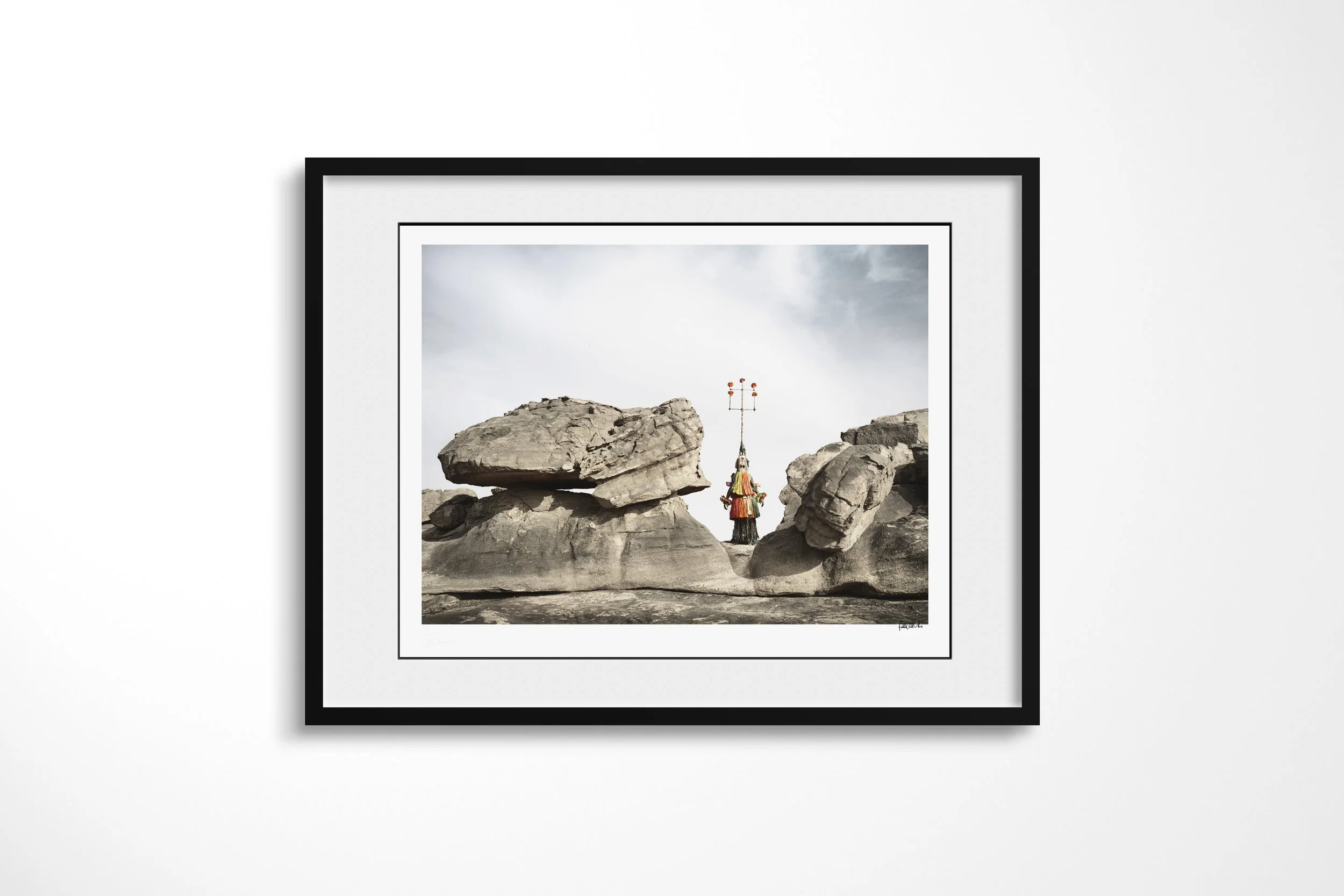 Framed presentation of Karamagra (Mask) in a black frame and white mount, showing a tall Dogon Karamagra mask standing between massive boulders.