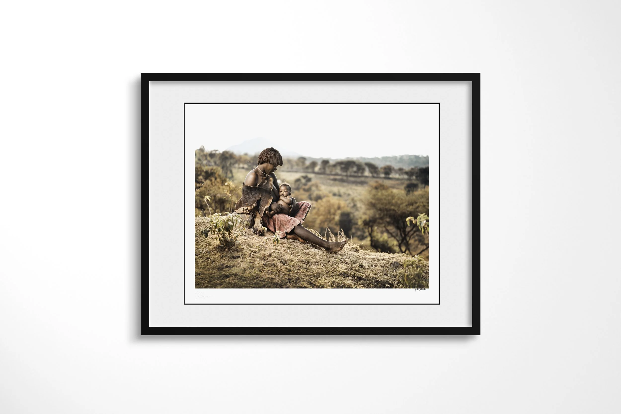 Framed presentation of Bami & Elsa in a black frame and white mount, showing a Banna mother seated on a hillside holding her young daughter across her lap.