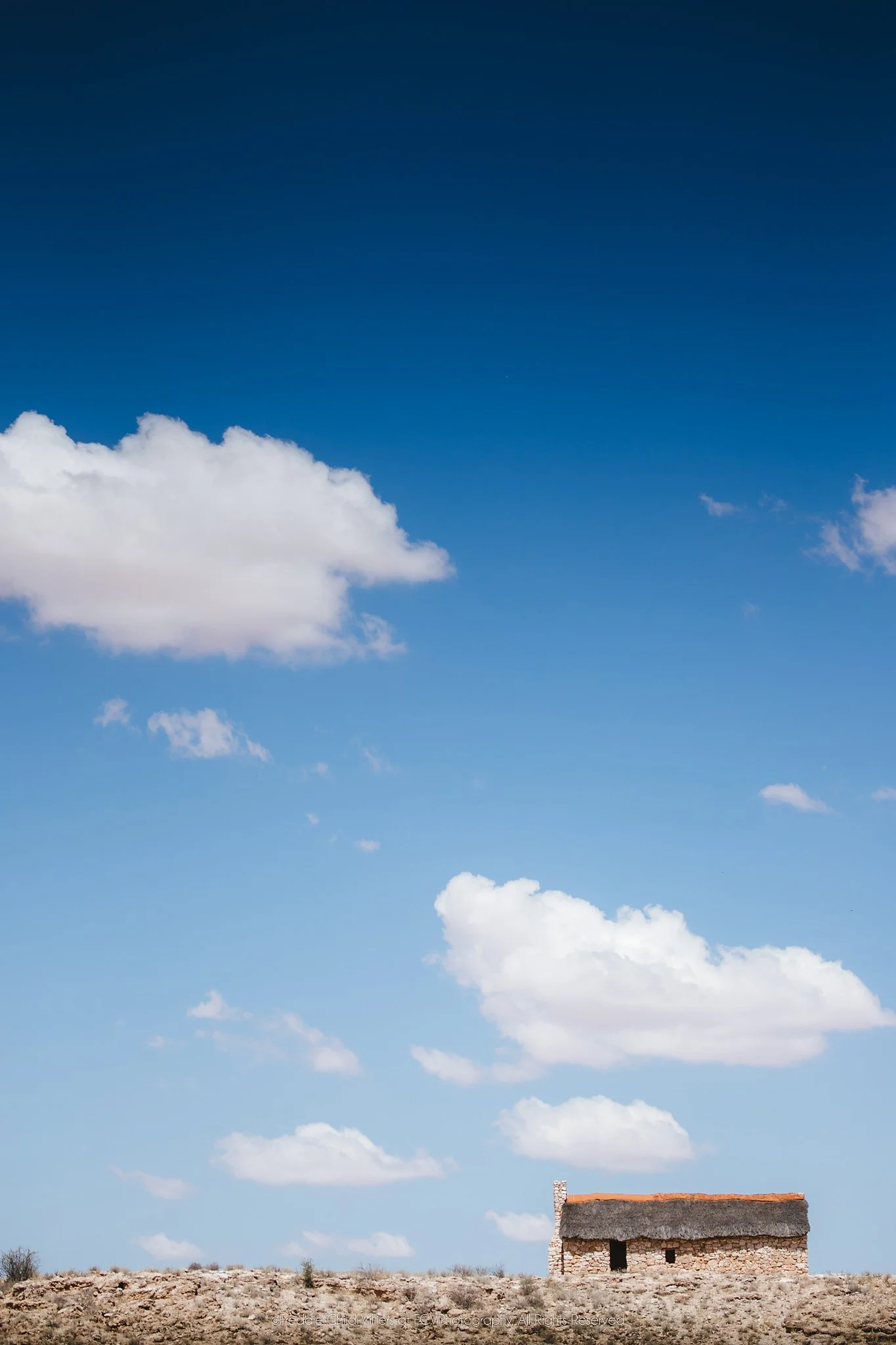 Small thatched house isolated on a barren ridge beneath a vast blue sky and scattered white clouds.