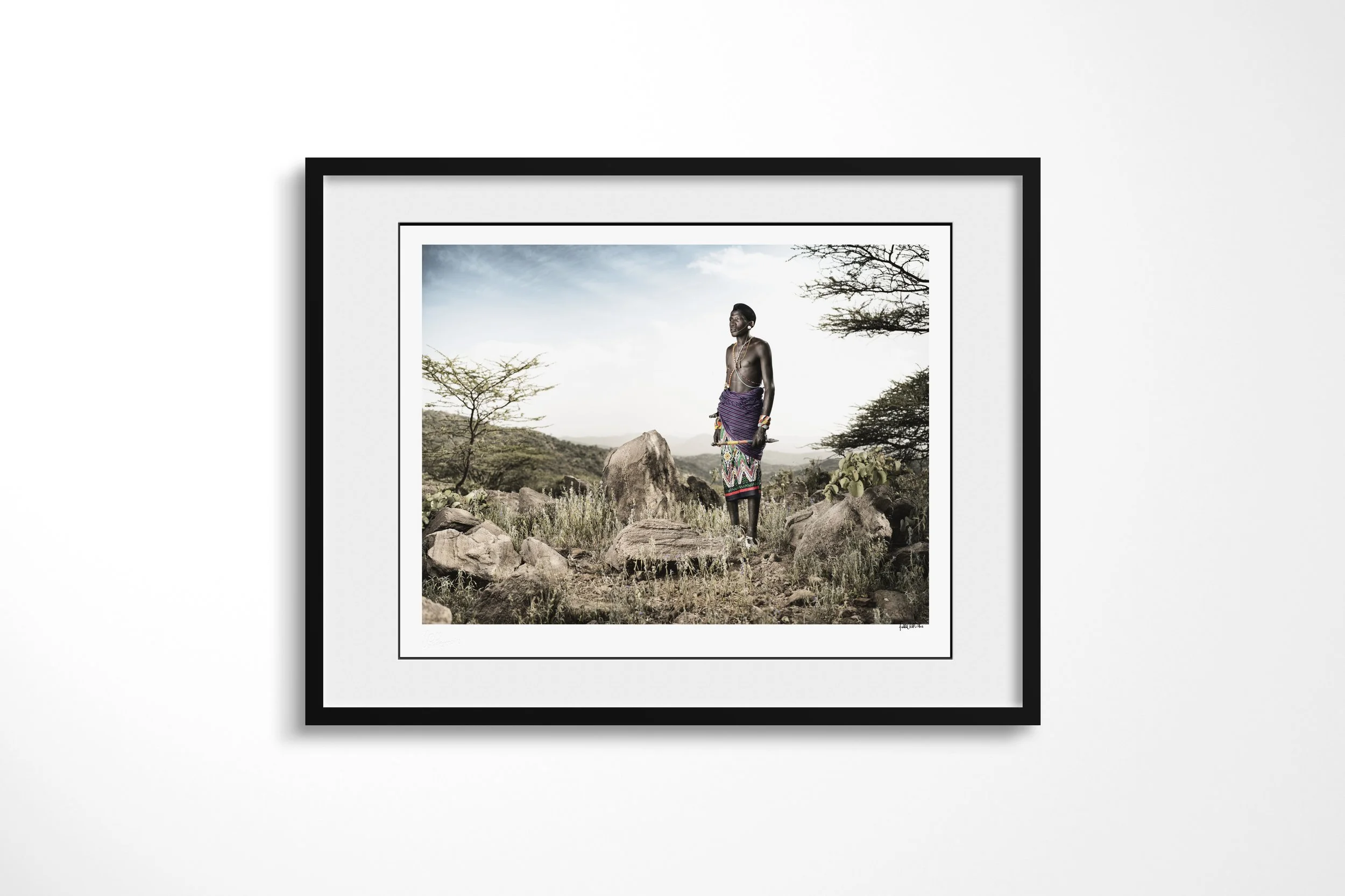 Framed presentation of Lkatinga in a black frame and white mount, showing a Samburu man in purple and patterned clothing standing among rocks in the Matthews Mountains.