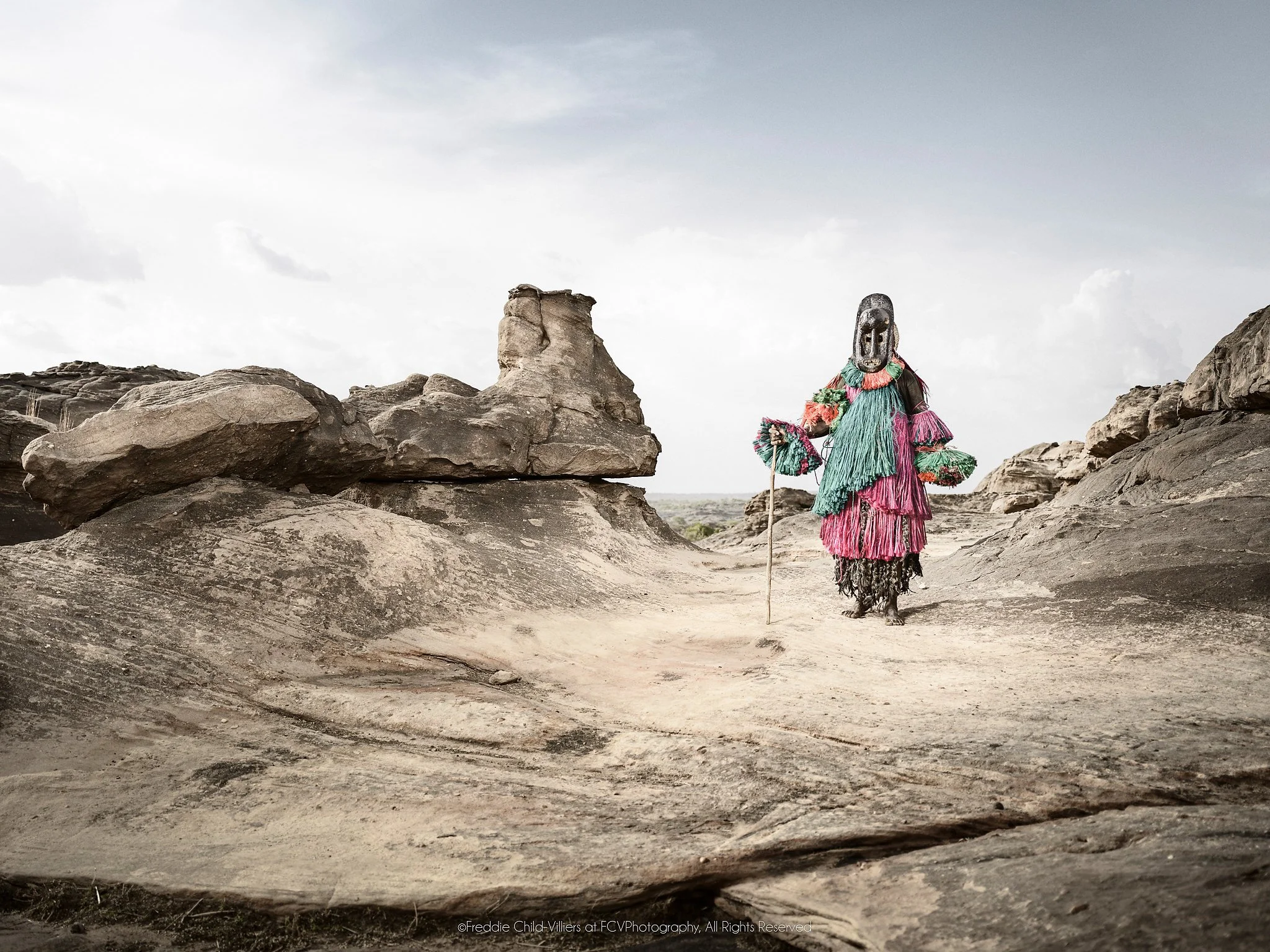 Dana (Mask), a Dogon hunter mask walking across open rock beneath a pale sky.