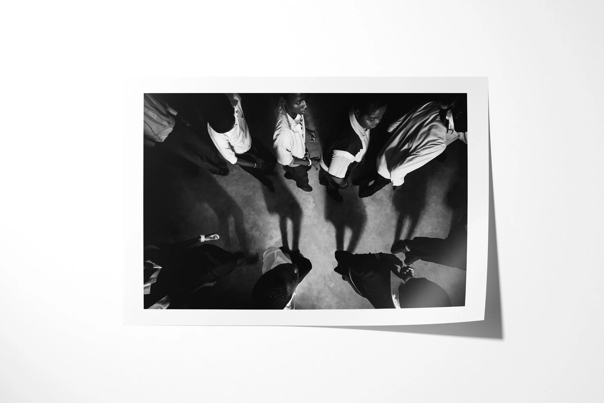 Unframed fine art paper print of Chant, showing a group of people seen from directly overhead, standing in a loose circle with long shadows across a concrete floor.