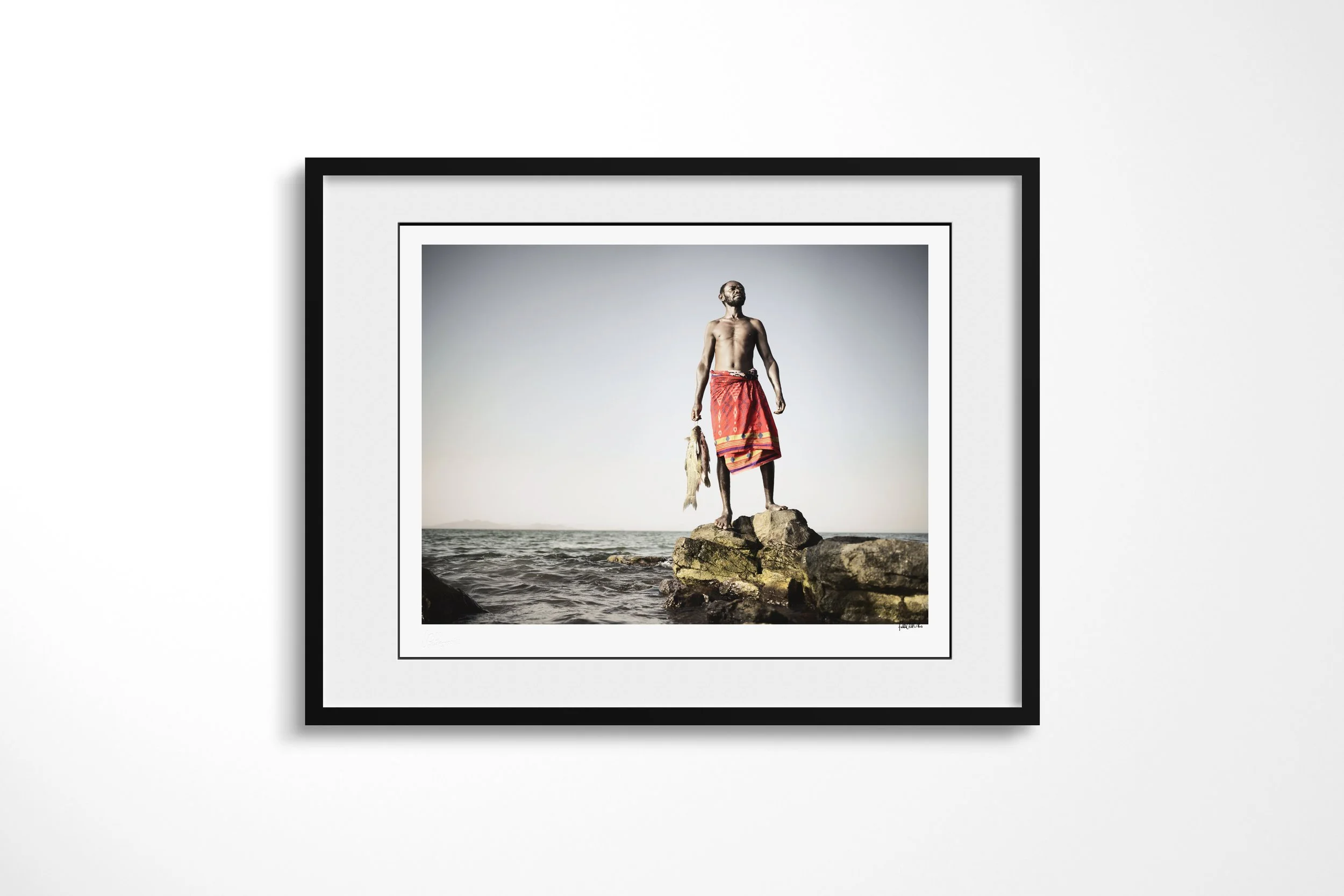 Framed presentation of Zacheus in a black frame and white mount, showing an El Molo fisherman standing on lakeside rocks holding two fish.