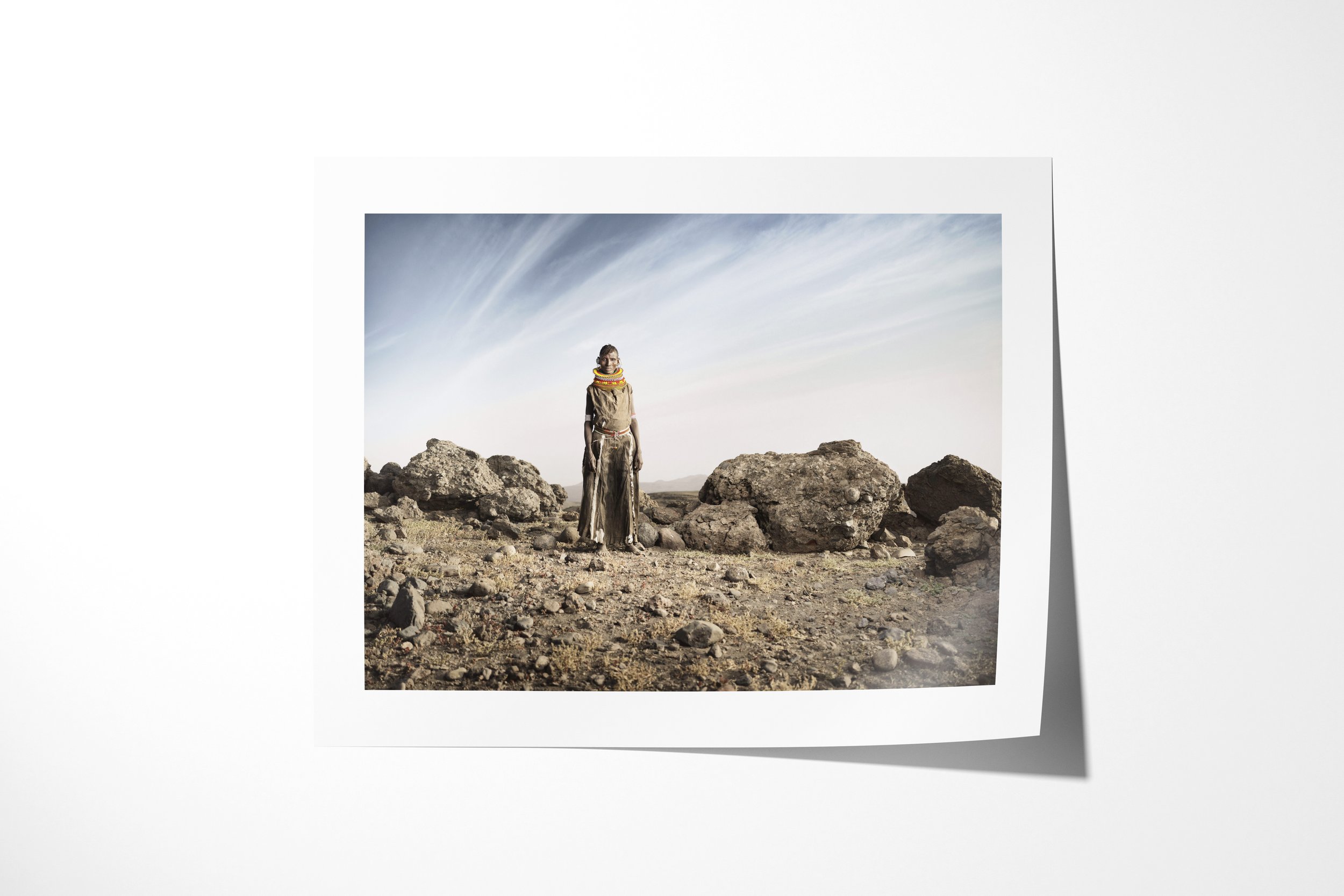 Fine art paper print of Aseguan, showing a Turkana woman in a long hide dress and beadwork standing among dark rocks in an arid landscape.