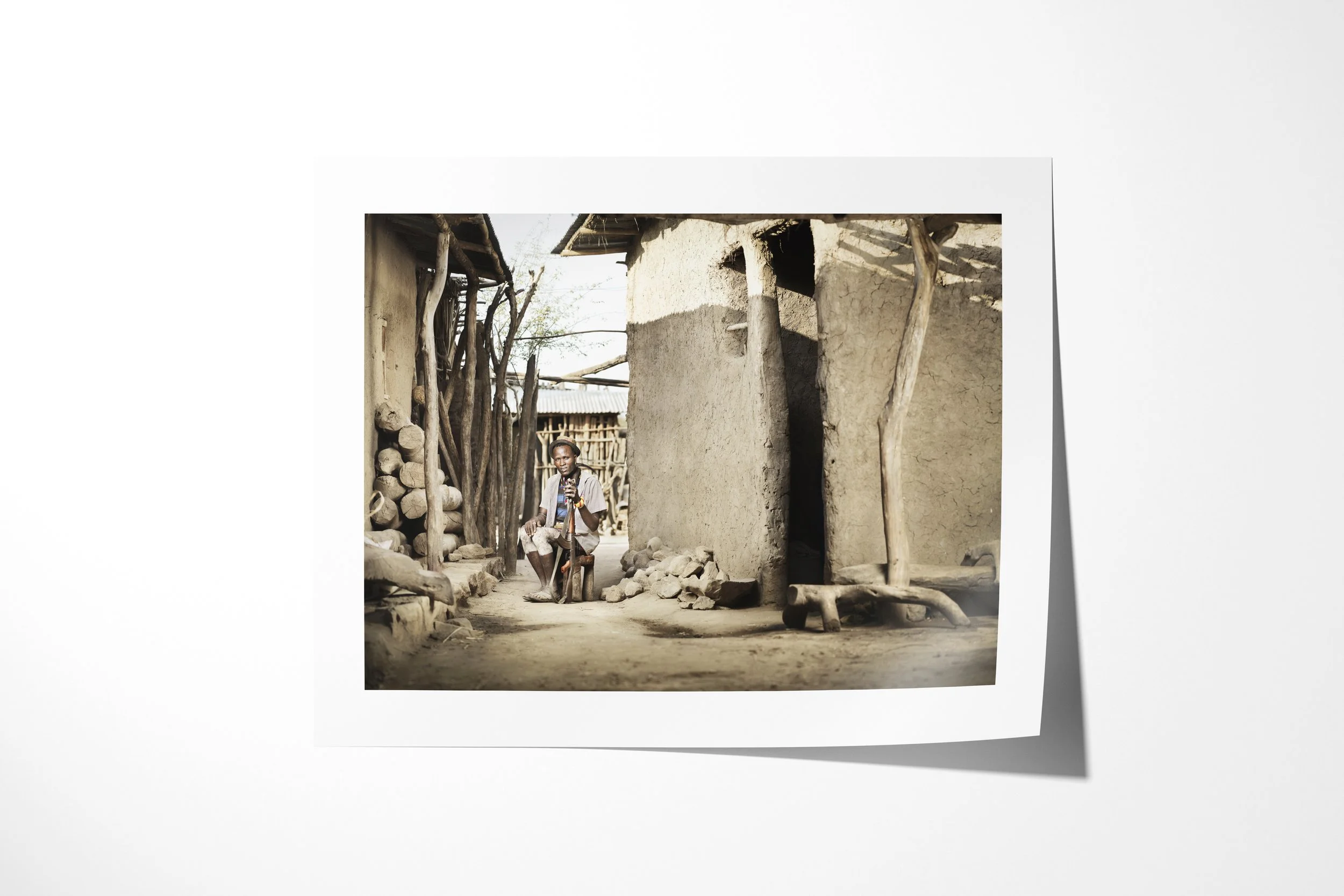 Fine art paper print of Bule, showing an Arbore man seated between earthen buildings with a staff and pale-painted legs.