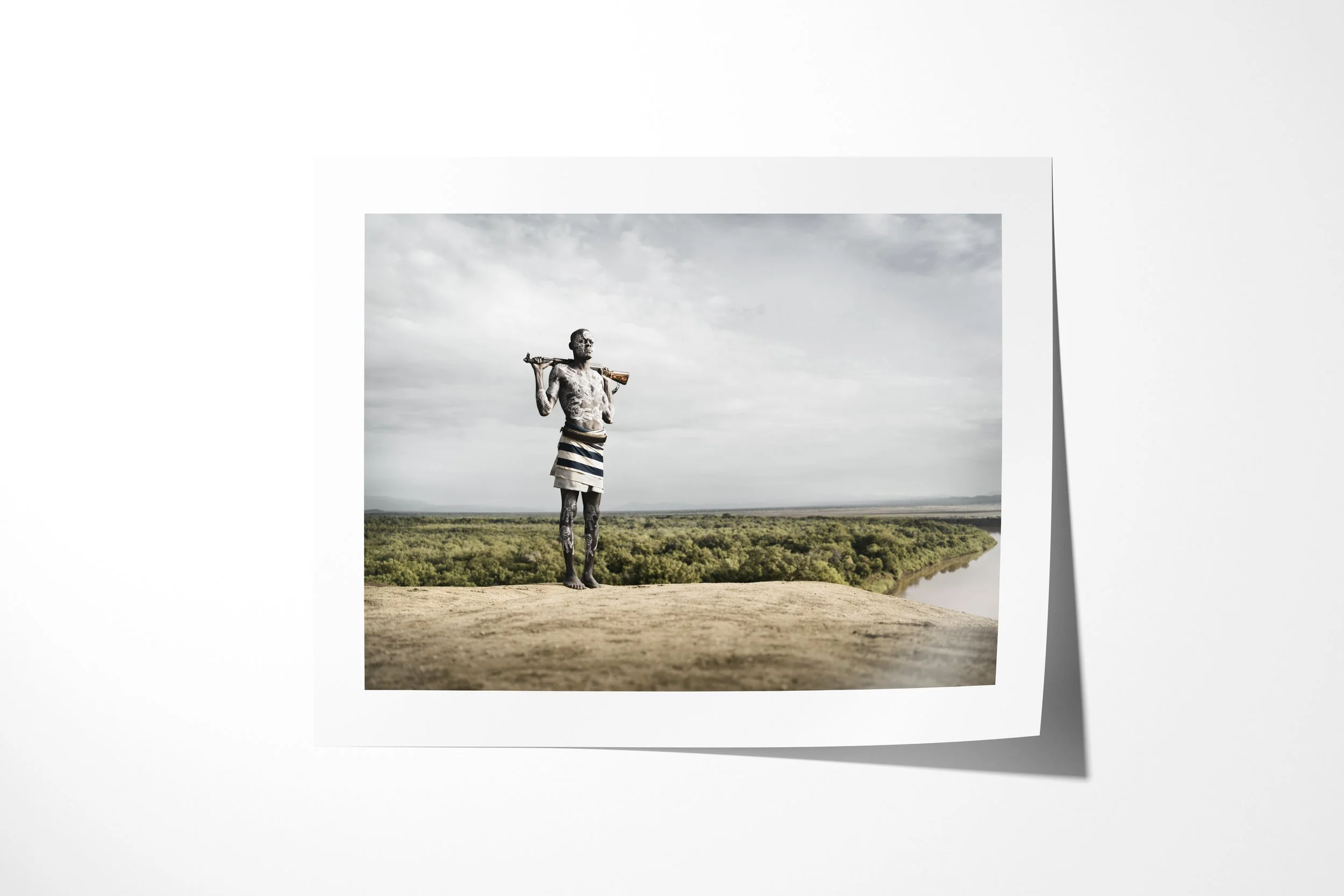Fine art paper print of Muda, showing a Kara man with painted skin and a rifle across his shoulders overlooking the Omo landscape.