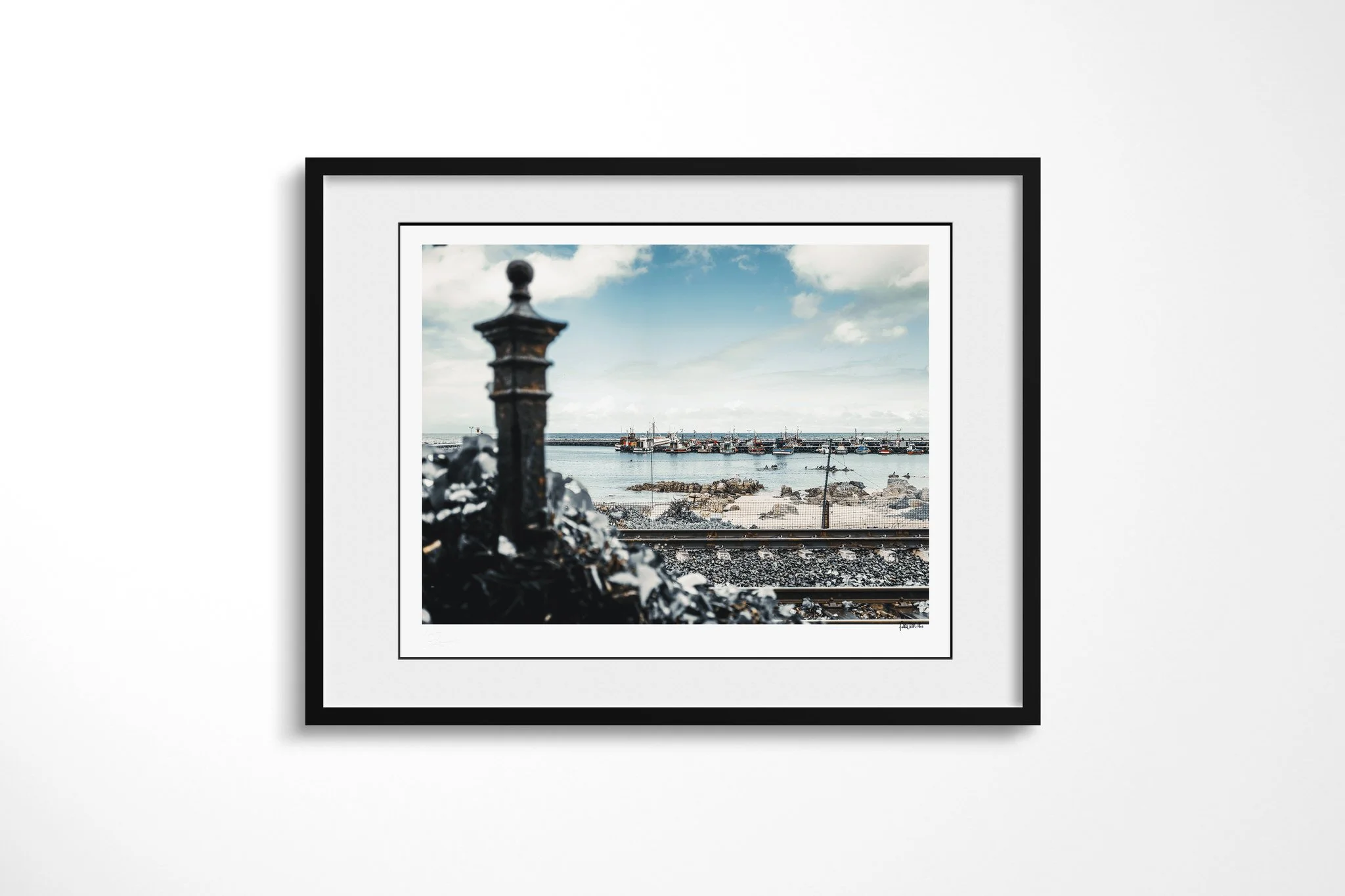 Black-framed fine art print of Harbour with a white mount, showing fishing boats moored across a calm harbour beyond a rocky shore and railway tracks, with a blurred iron post in front.