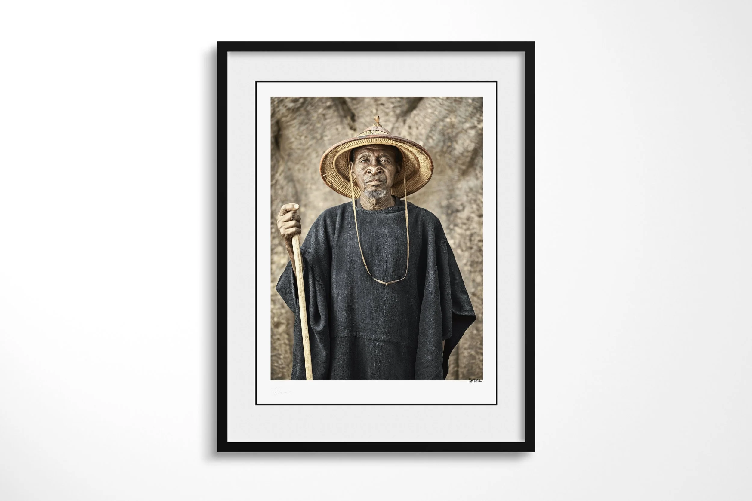 Framed presentation of Tigué in a black frame and white mount, showing Tigué in a dark robe and broad straw hat holding a staff before a baobab trunk.