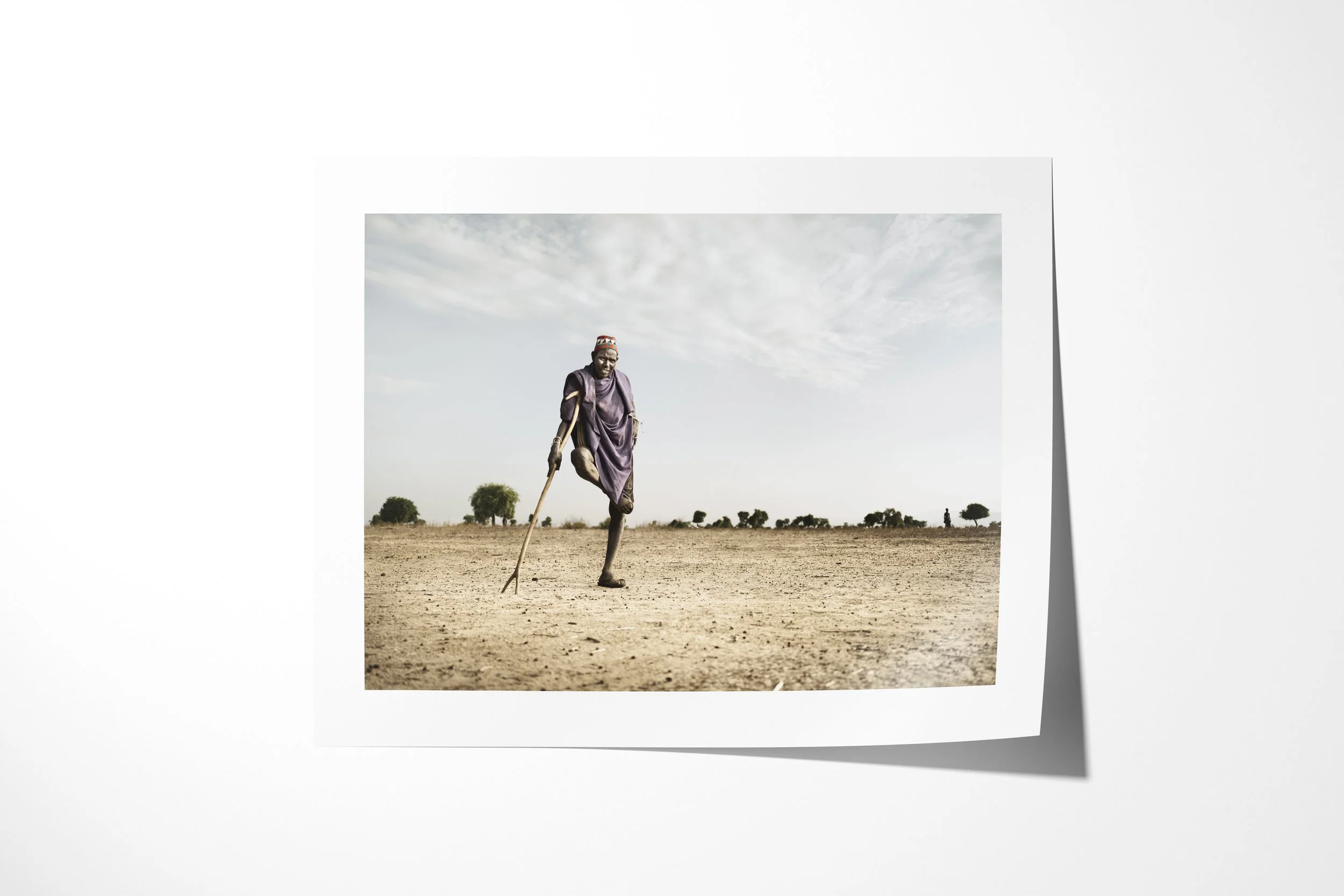 Fine art paper print of Buba, showing an Arbore man balancing on one leg while leaning on a long staff in an arid landscape.