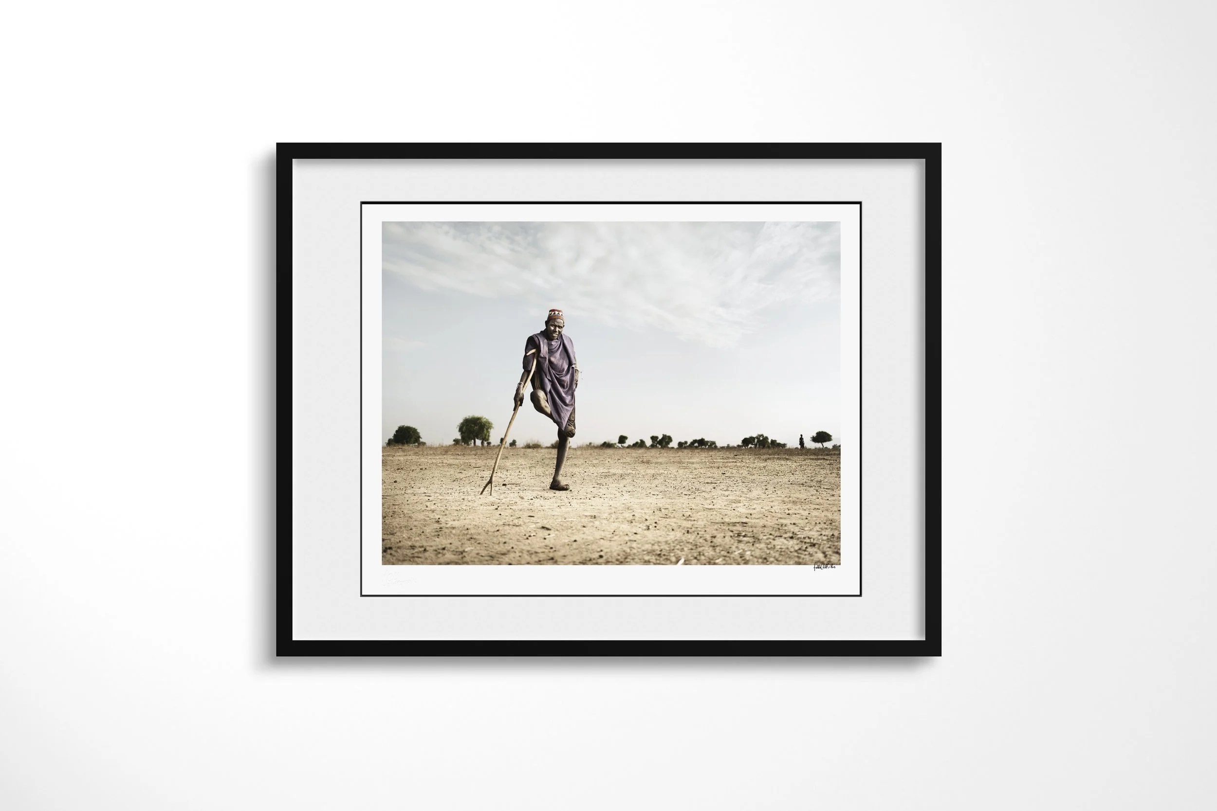 Framed presentation of Buba in a black frame and white mount, showing an Arbore man balancing on one leg while leaning on a long staff in an arid landscape.