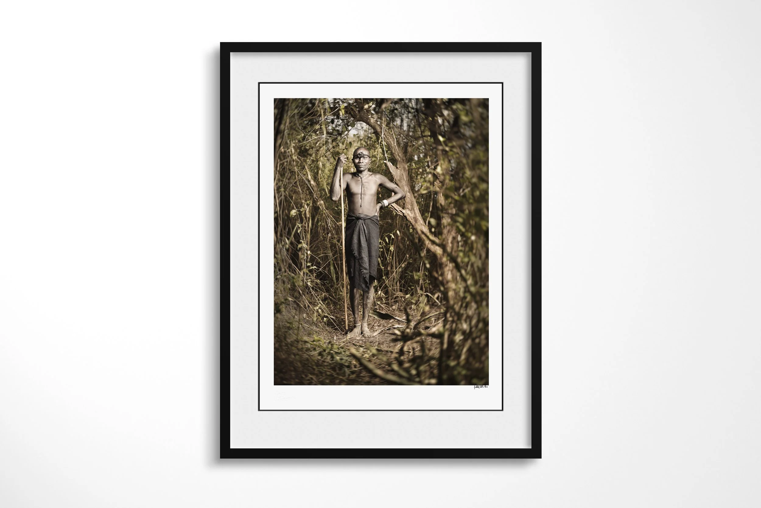 Framed presentation of Lemoi in a black frame and white mount, showing a Mursi man holding a long staff on a narrow path through dense woodland.